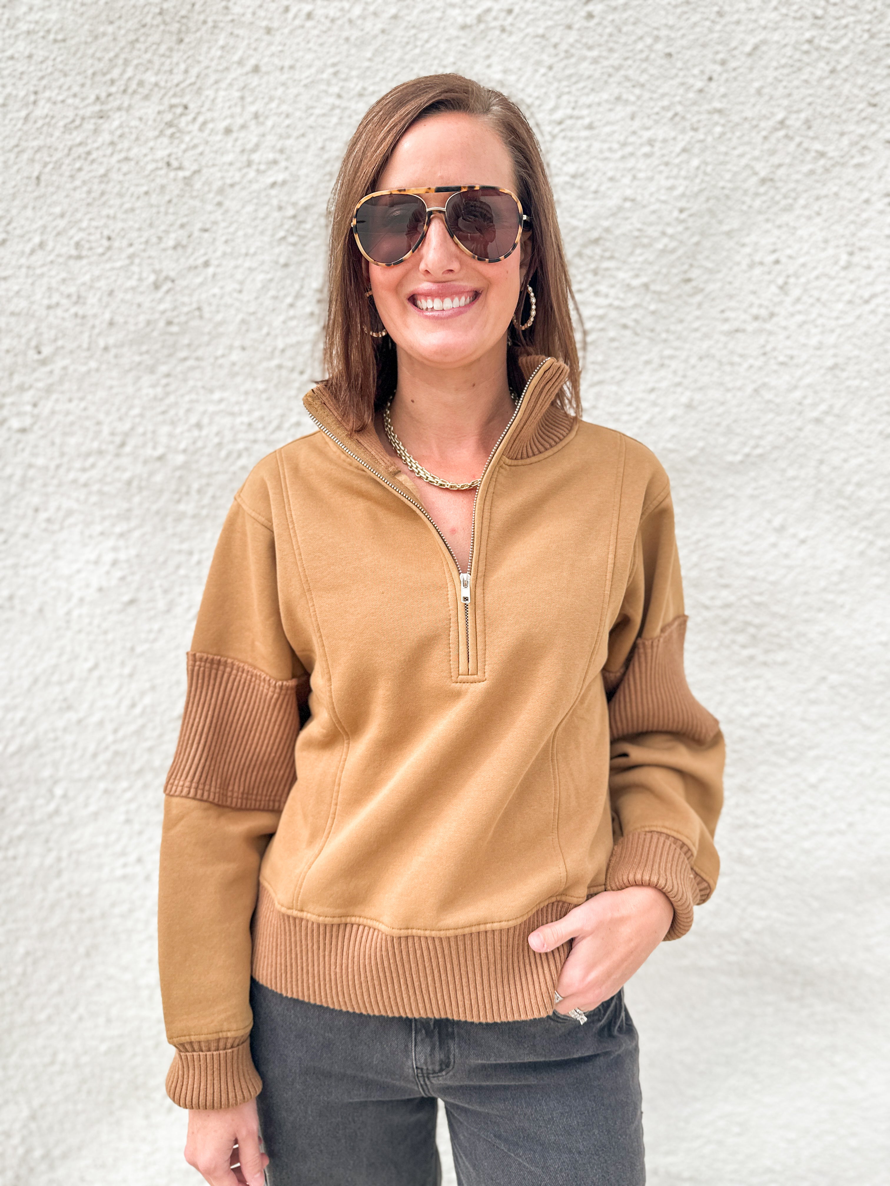 A woman wearing a tan quarter-zip pullover sweater with ribbed trim and darker brown ribbed elbow patches, paired with dark gray jeans, standing against a textured white wall.