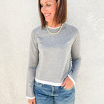 A woman wearing a light gray long-sleeve raglan sweatshirt with white ribbed trim at the collar, cuffs, and hem, paired with blue jeans, standing against a textured off-white wall.