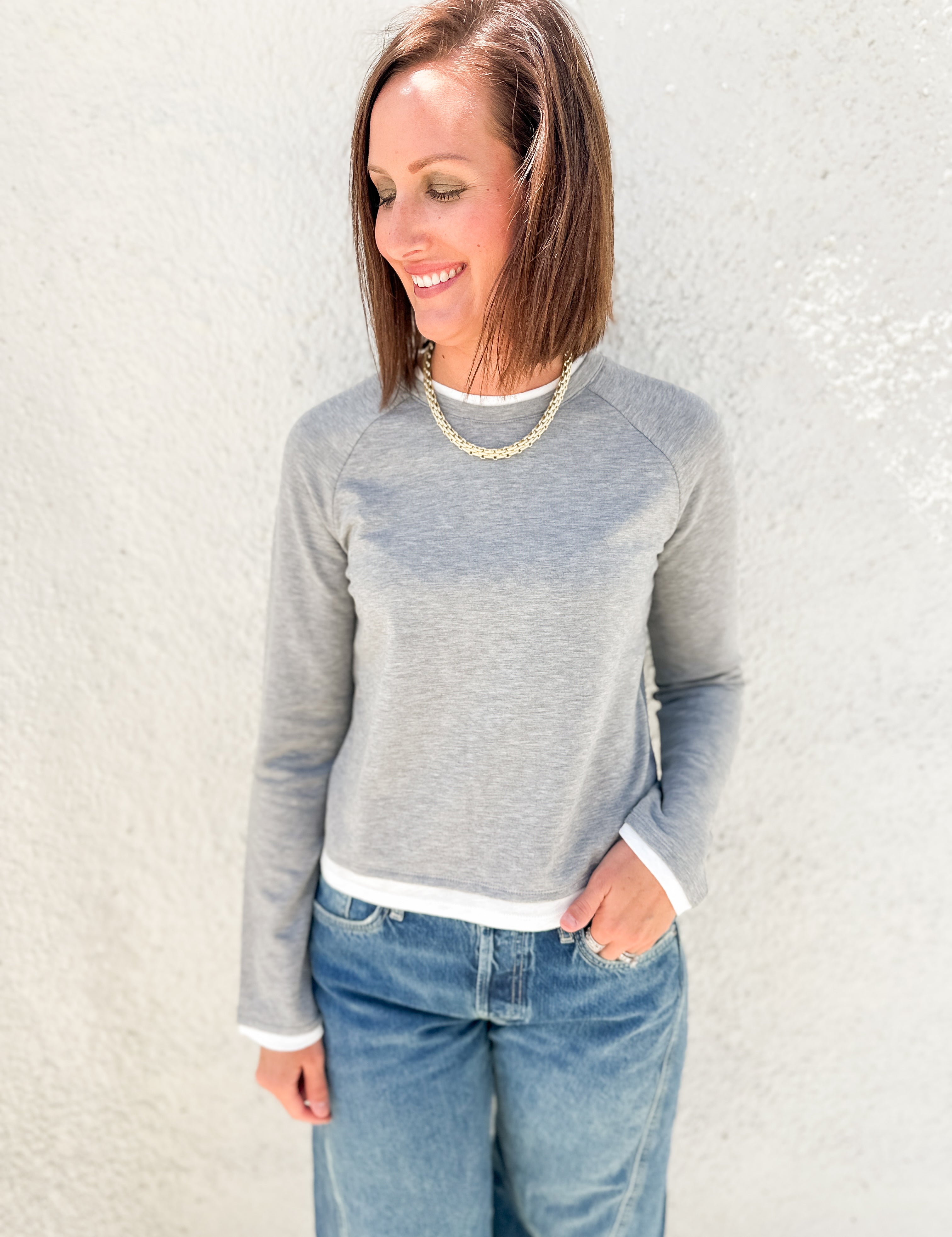 A woman wearing a light gray long-sleeve raglan sweatshirt with white ribbed trim at the collar, cuffs, and hem, paired with blue jeans, standing against a textured off-white wall.