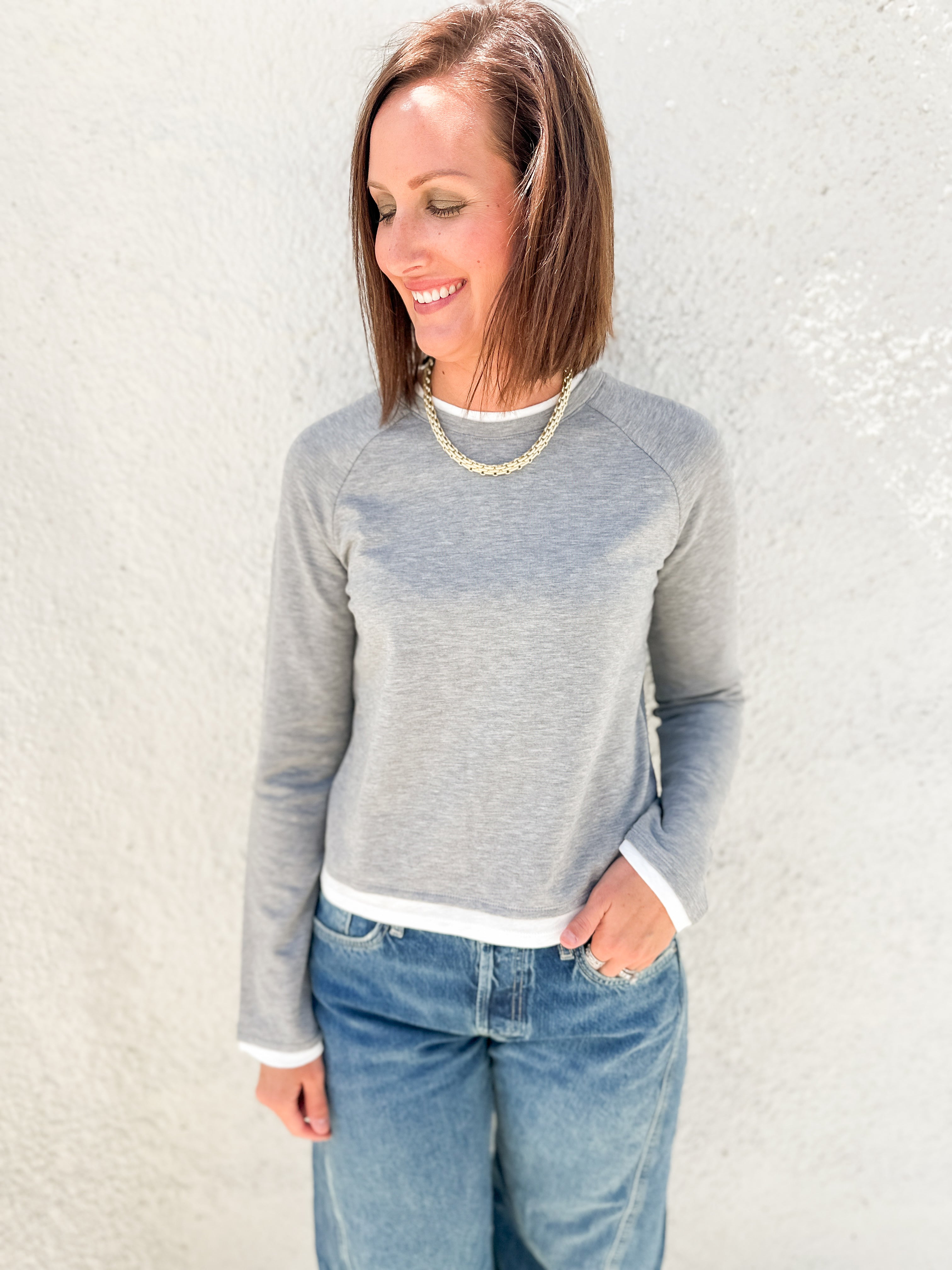 A woman wearing a light gray long-sleeve raglan sweatshirt with white ribbed trim at the collar, cuffs, and hem, paired with blue jeans, standing against a textured off-white wall.