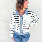 A woman wearing a white and black horizontally striped knit cardigan over a light blue denim button-up shirt and blue jeans, standing against a white stucco wall.