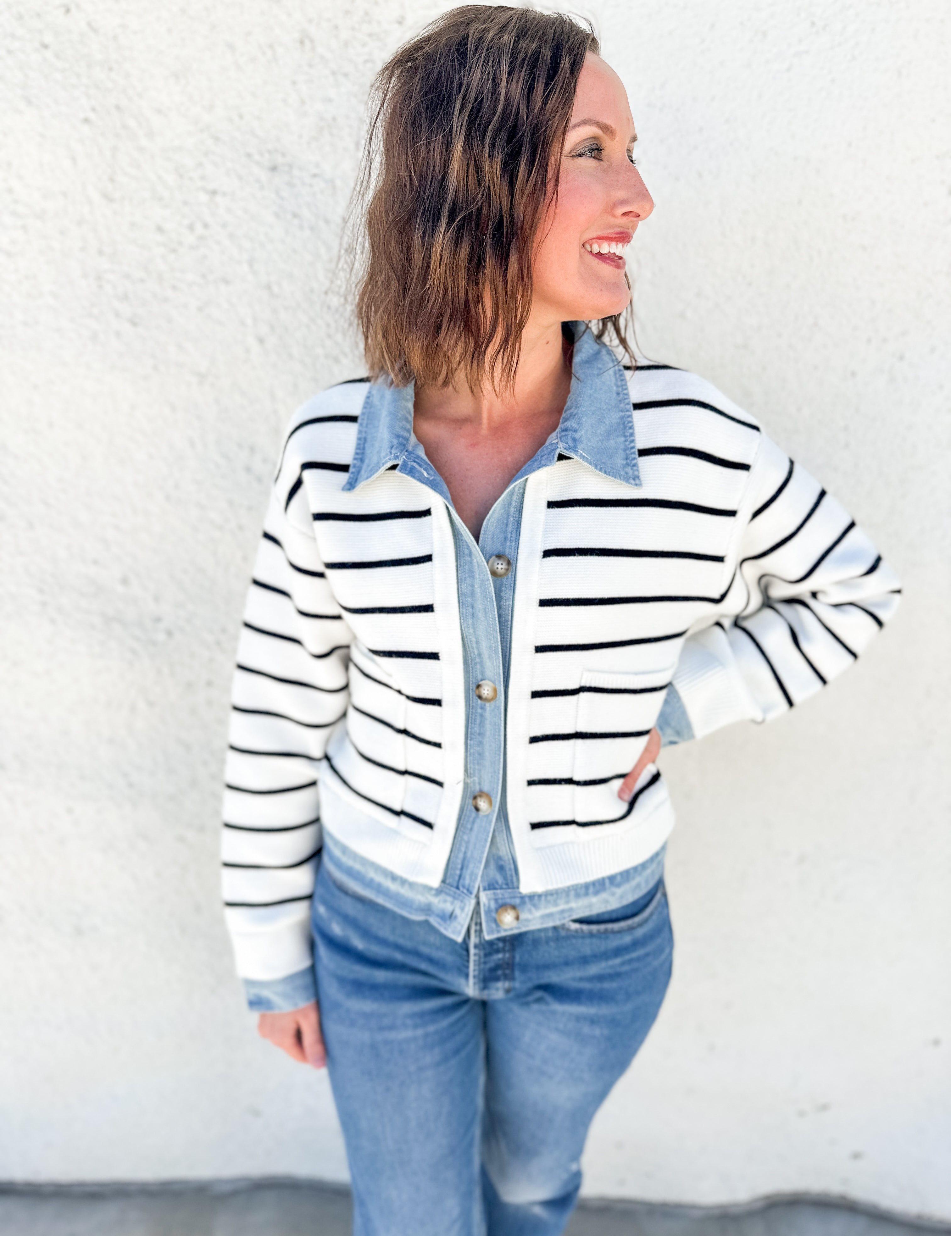 A woman wearing a white and black horizontally striped knit cardigan over a light blue denim button-up shirt and blue jeans, standing against a white stucco wall.