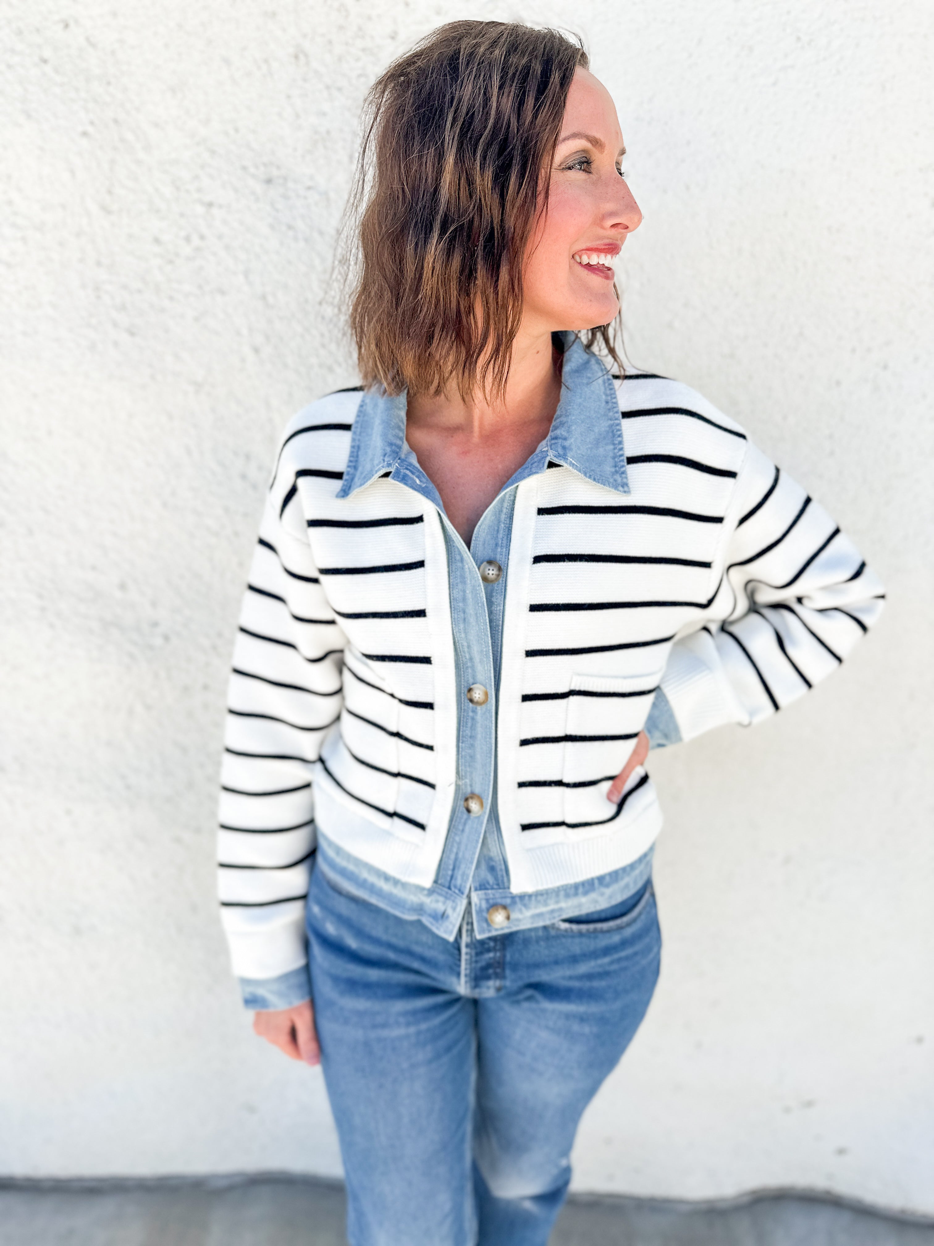 A woman wearing a white and black horizontally striped knit cardigan over a light blue denim button-up shirt and blue jeans, standing against a white stucco wall.