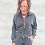 A woman wearing a matching charcoal gray funnel neck zip-up sweatshirt and barrel sweatpants set, standing against a textured white wall.