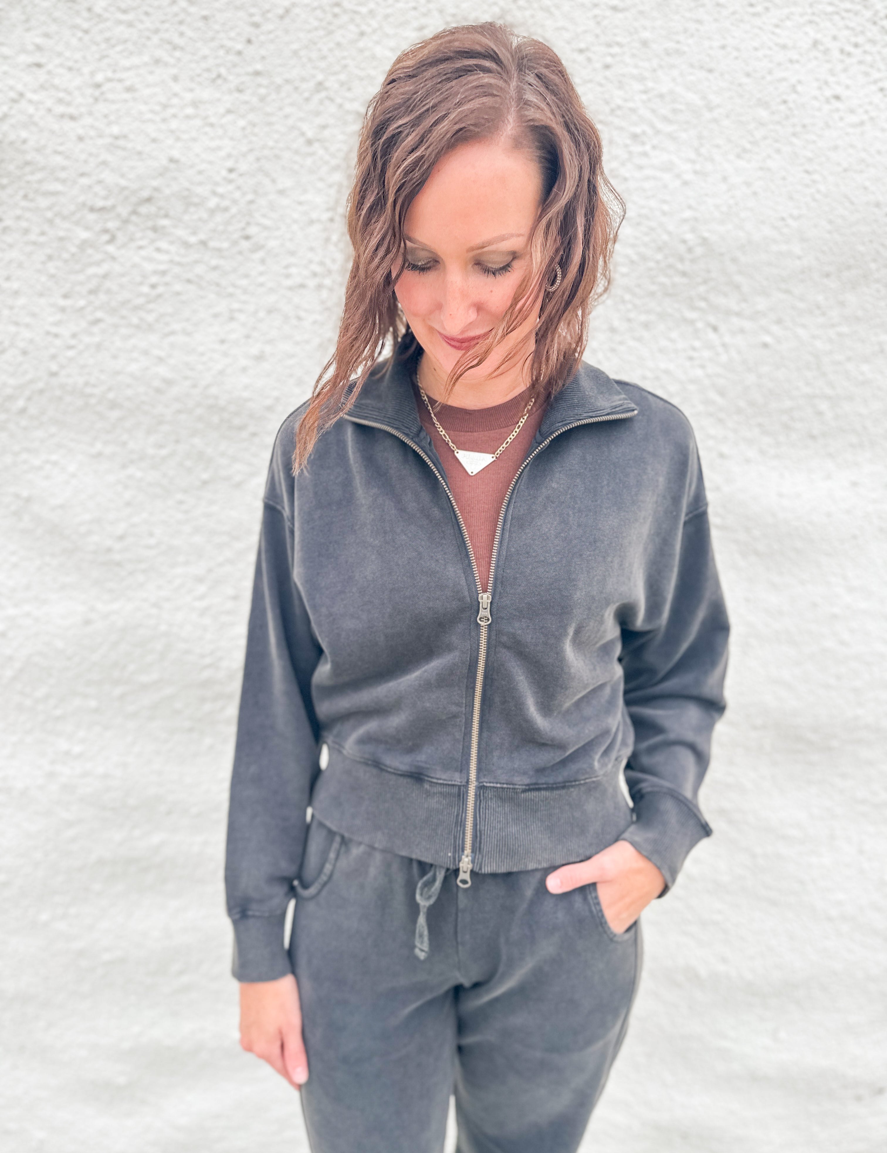 A woman wearing a matching charcoal gray funnel neck zip-up sweatshirt and barrel sweatpants set, standing against a textured white wall.