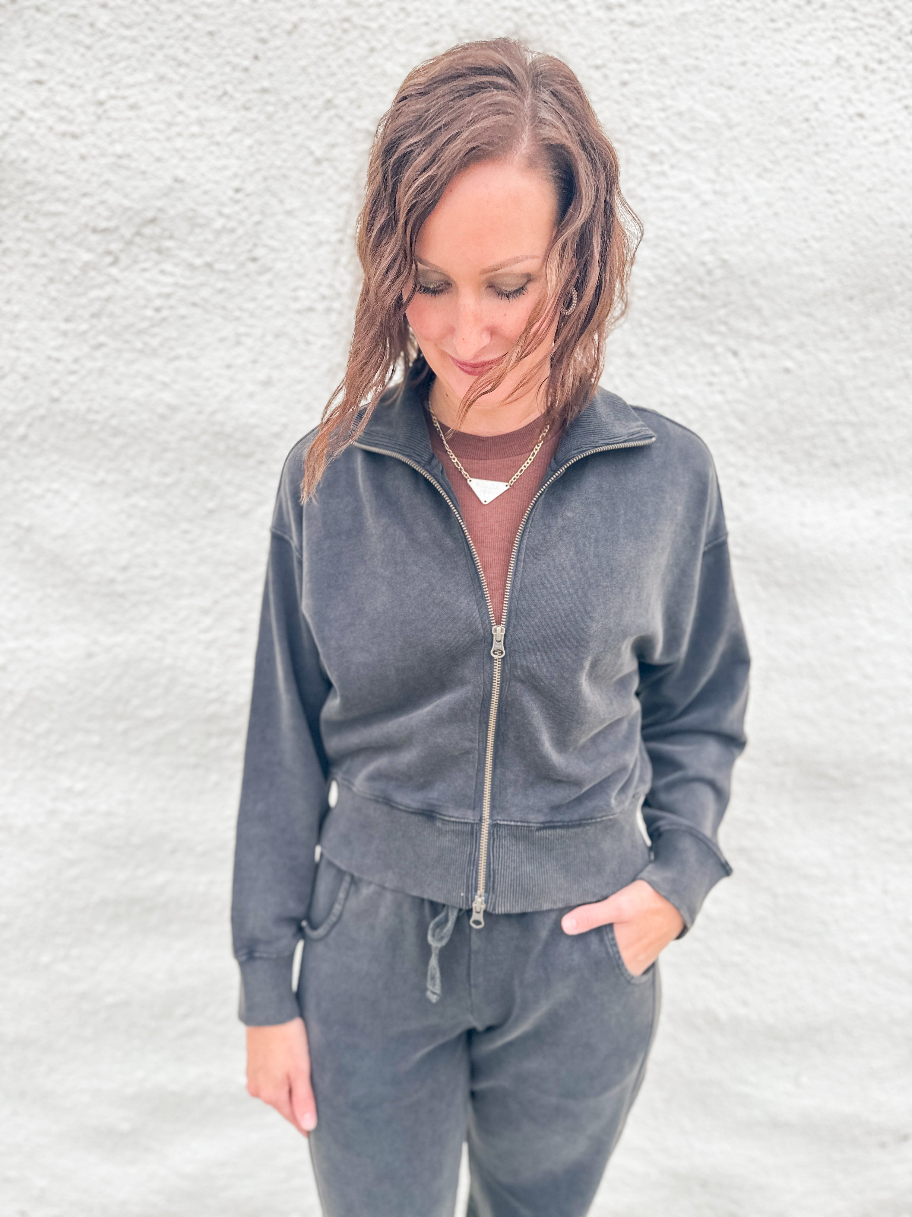 A woman wearing a matching charcoal gray funnel neck zip-up sweatshirt and barrel sweatpants set, standing against a textured white wall.