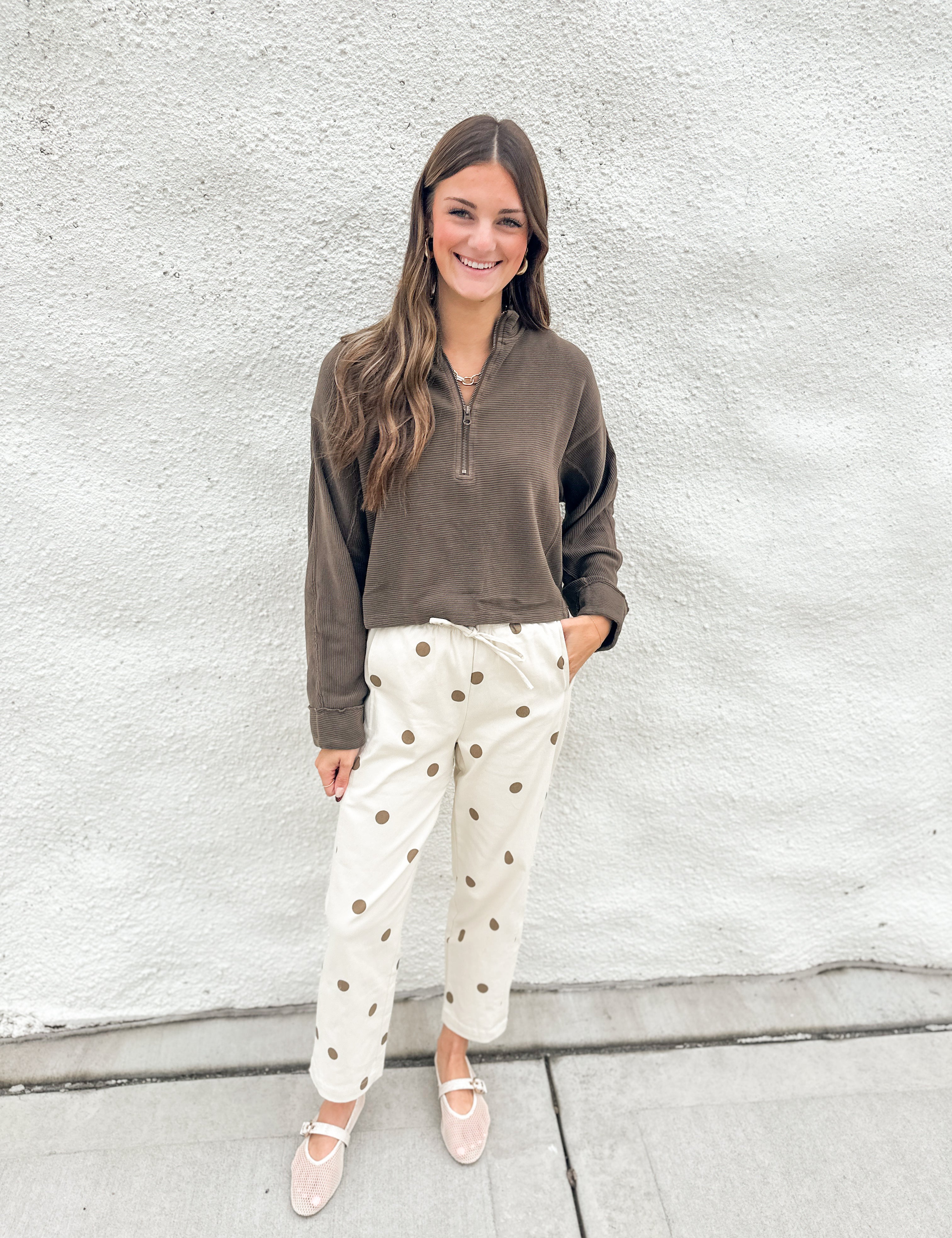 A woman stands against a white textured wall wearing a brown quarter-zip long-sleeve top, cream pants with brown polka dots, and cream mesh mary jane flats.