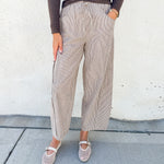 A person wearing loose, wide-leg cropped pants in a light taupe/beige textured fabric with a drawstring waist, paired with a dark brown long-sleeve top and beige Mary Jane shoes, standing against a white stucco wall.
