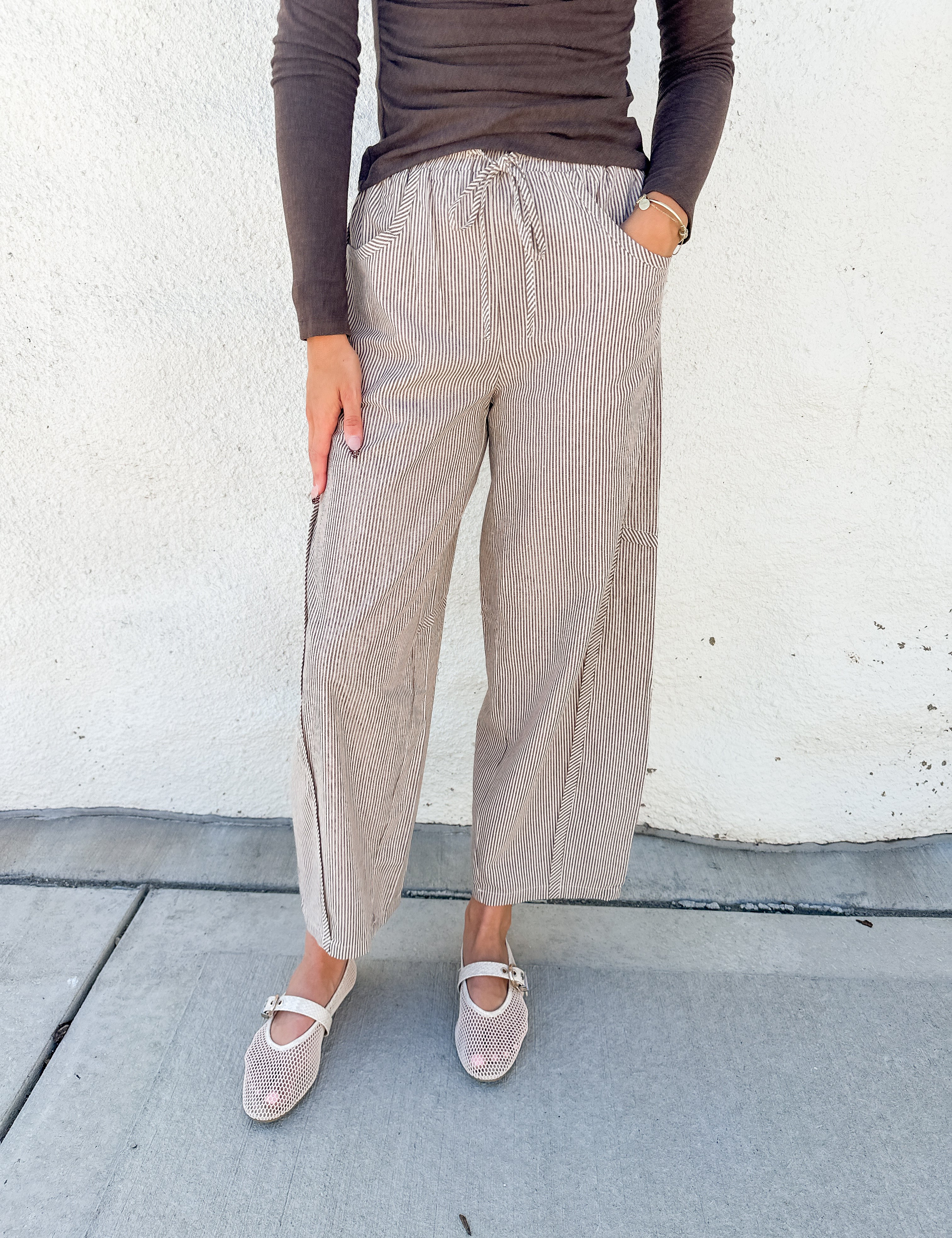 A person wearing loose, wide-leg cropped pants in a light taupe/beige textured fabric with a drawstring waist, paired with a dark brown long-sleeve top and beige Mary Jane shoes, standing against a white stucco wall.
