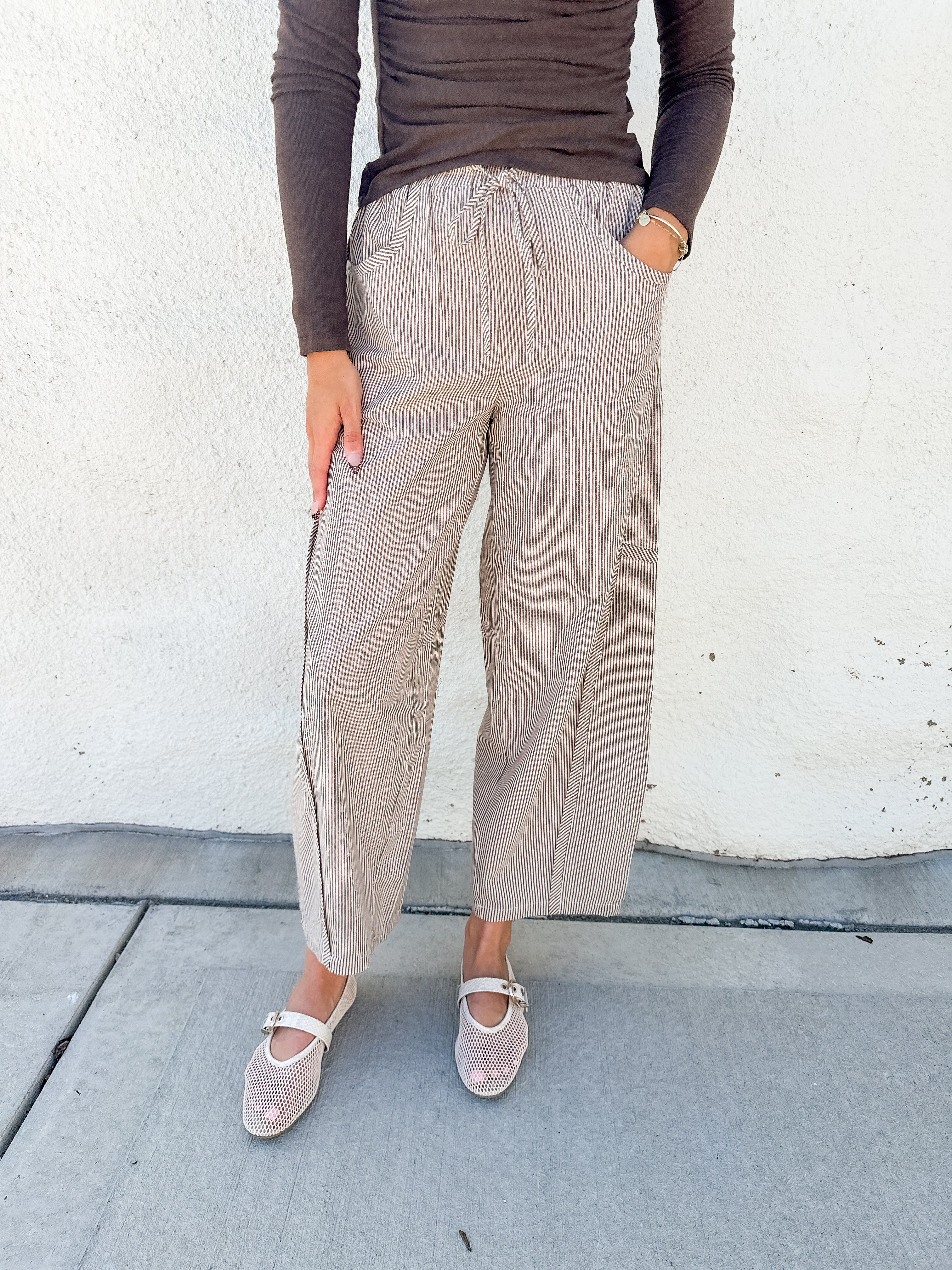 A person wearing loose, wide-leg cropped pants in a light taupe/beige textured fabric with a drawstring waist, paired with a dark brown long-sleeve top and beige Mary Jane shoes, standing against a white stucco wall.