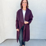 A woman modeling a long, open-front wrap coat in a deep plum purple color, worn over a white top and black pants, standing against a textured off-white wall.