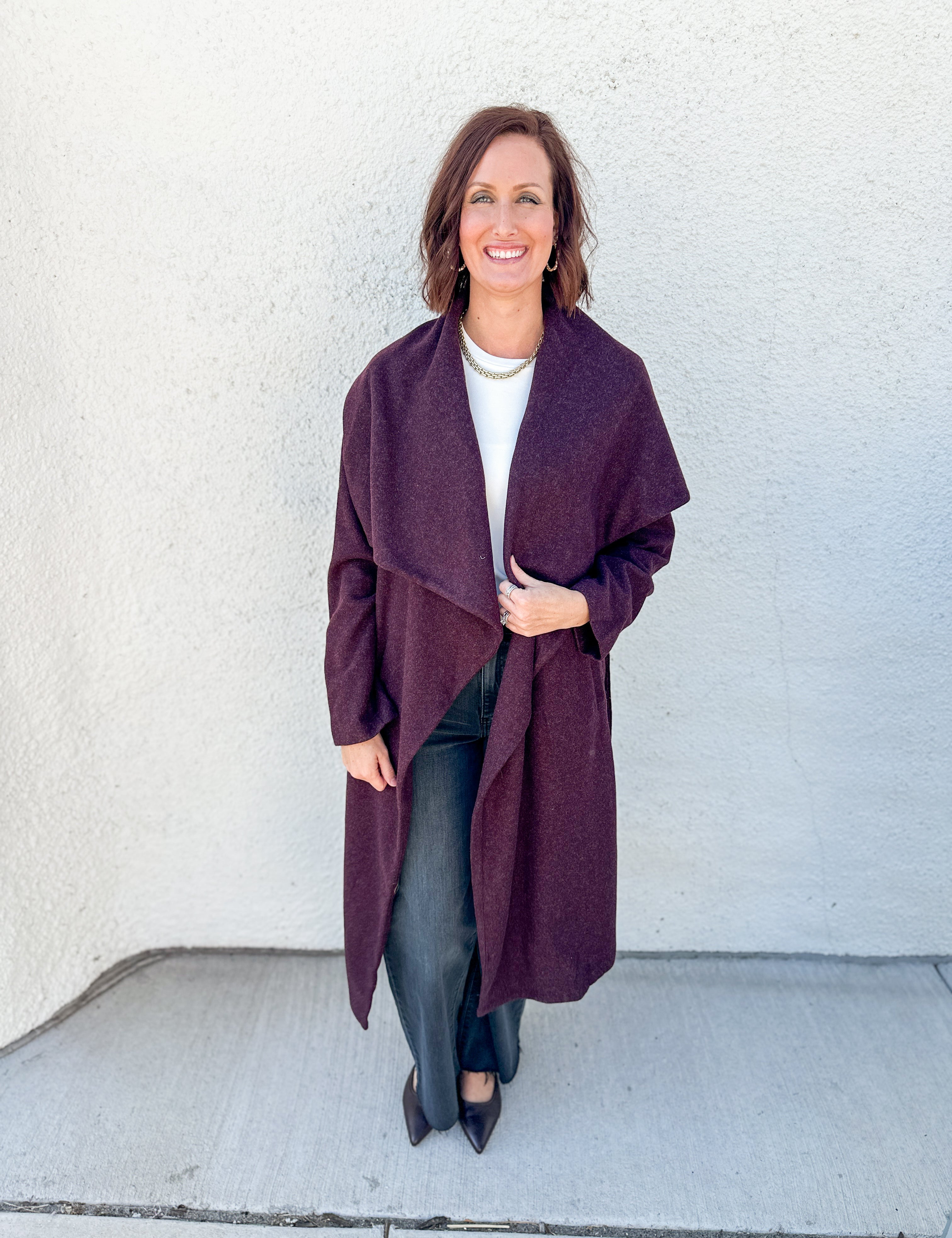A woman modeling a long, open-front wrap coat in a deep plum purple color, worn over a white top and black pants, standing against a textured off-white wall.