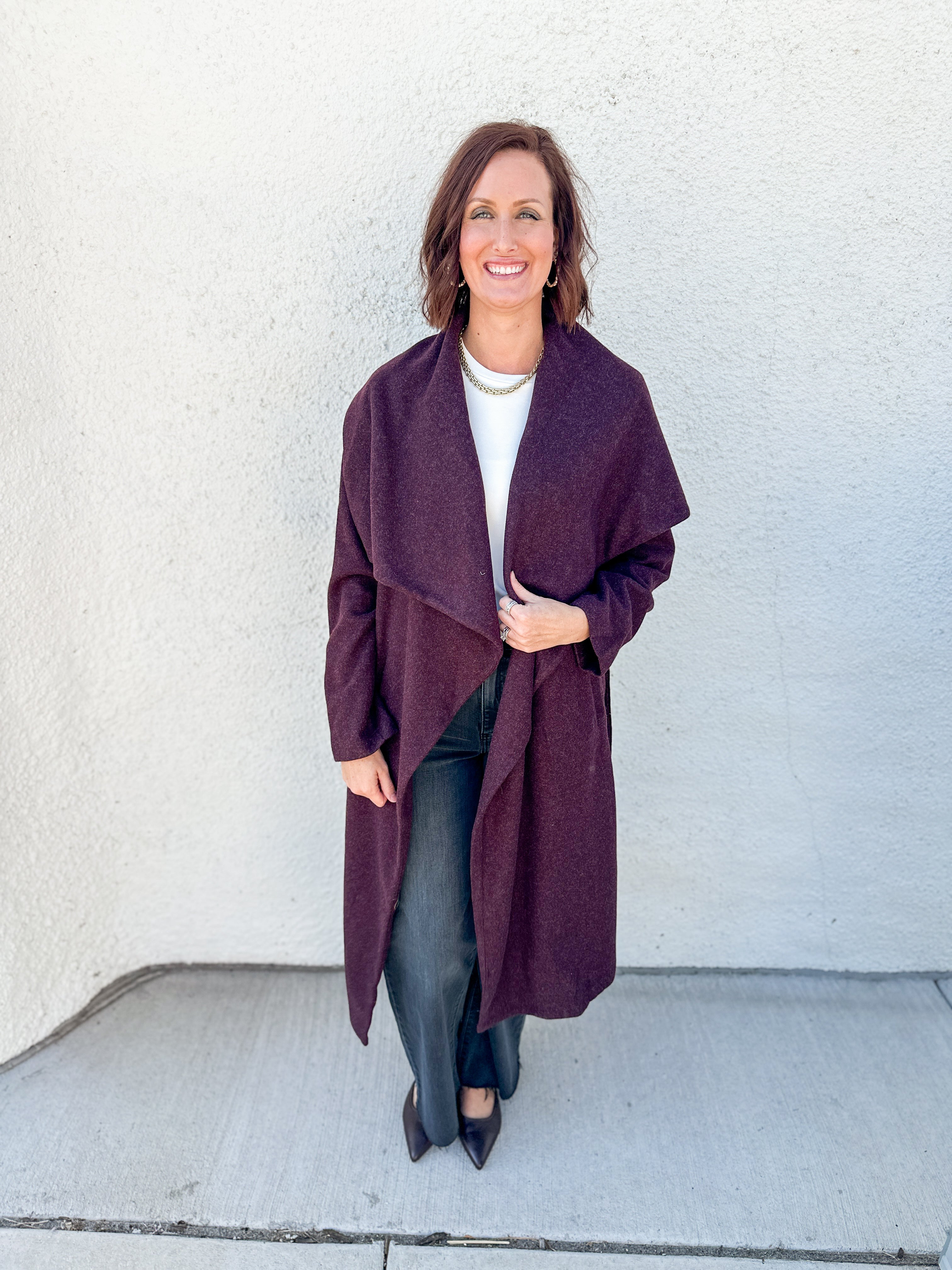 A woman modeling a long, open-front wrap coat in a deep plum purple color, worn over a white top and black pants, standing against a textured off-white wall.