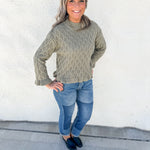 A smiling woman with gray hair stands outdoors against a white stucco wall, wearing a textured cable-knit sweater in a muted sage green, blue jeans, and dark loafers.