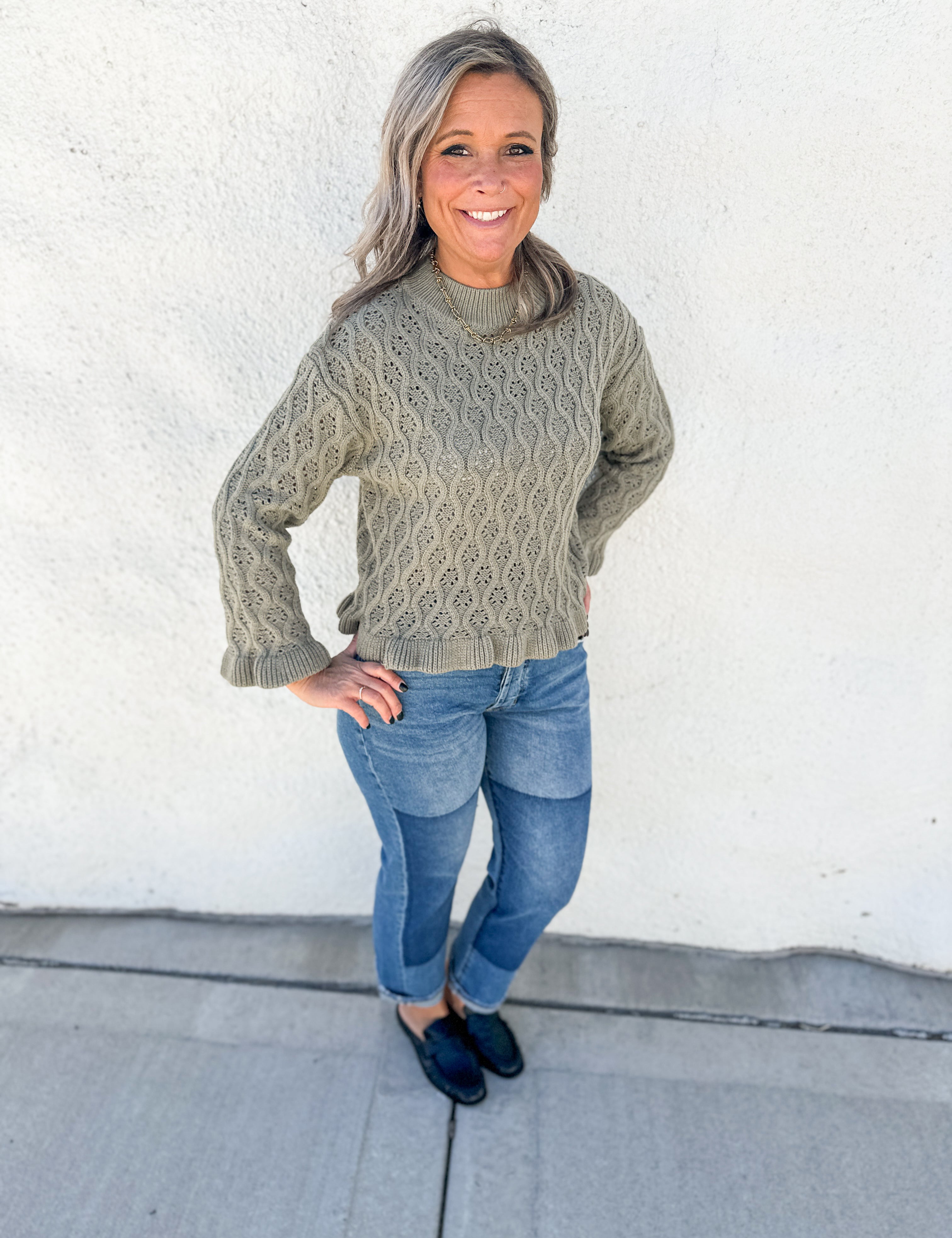 A smiling woman with gray hair stands outdoors against a white stucco wall, wearing a textured cable-knit sweater in a muted sage green, blue jeans, and dark loafers.