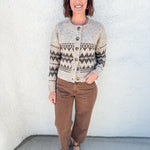 A woman stands against a white stucco wall wearing a cream and brown Fair Isle knit cardigan by Heartloom with brown barrel leg pants and mahogany brown pointed-toe flats.
