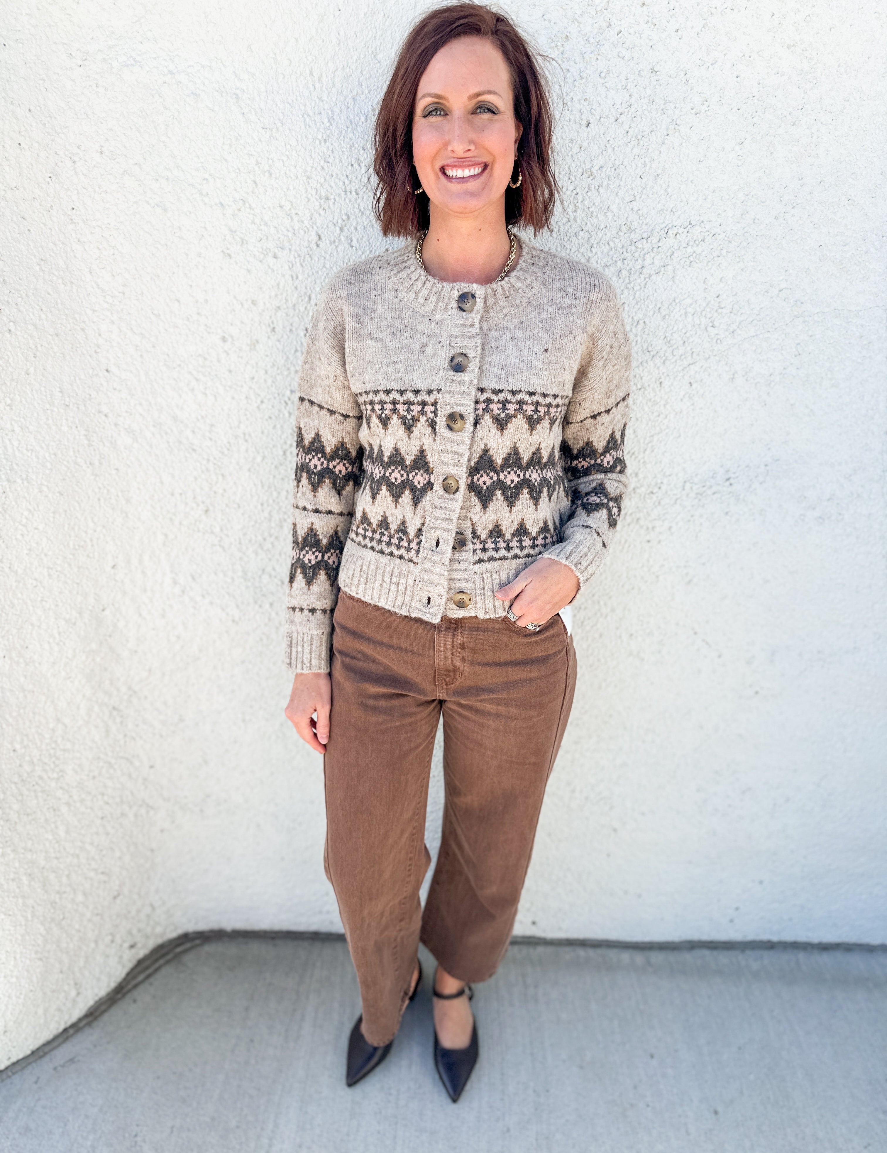 A woman stands against a white stucco wall wearing a cream and brown Fair Isle knit cardigan by Heartloom with brown barrel leg pants and mahogany brown pointed-toe flats.