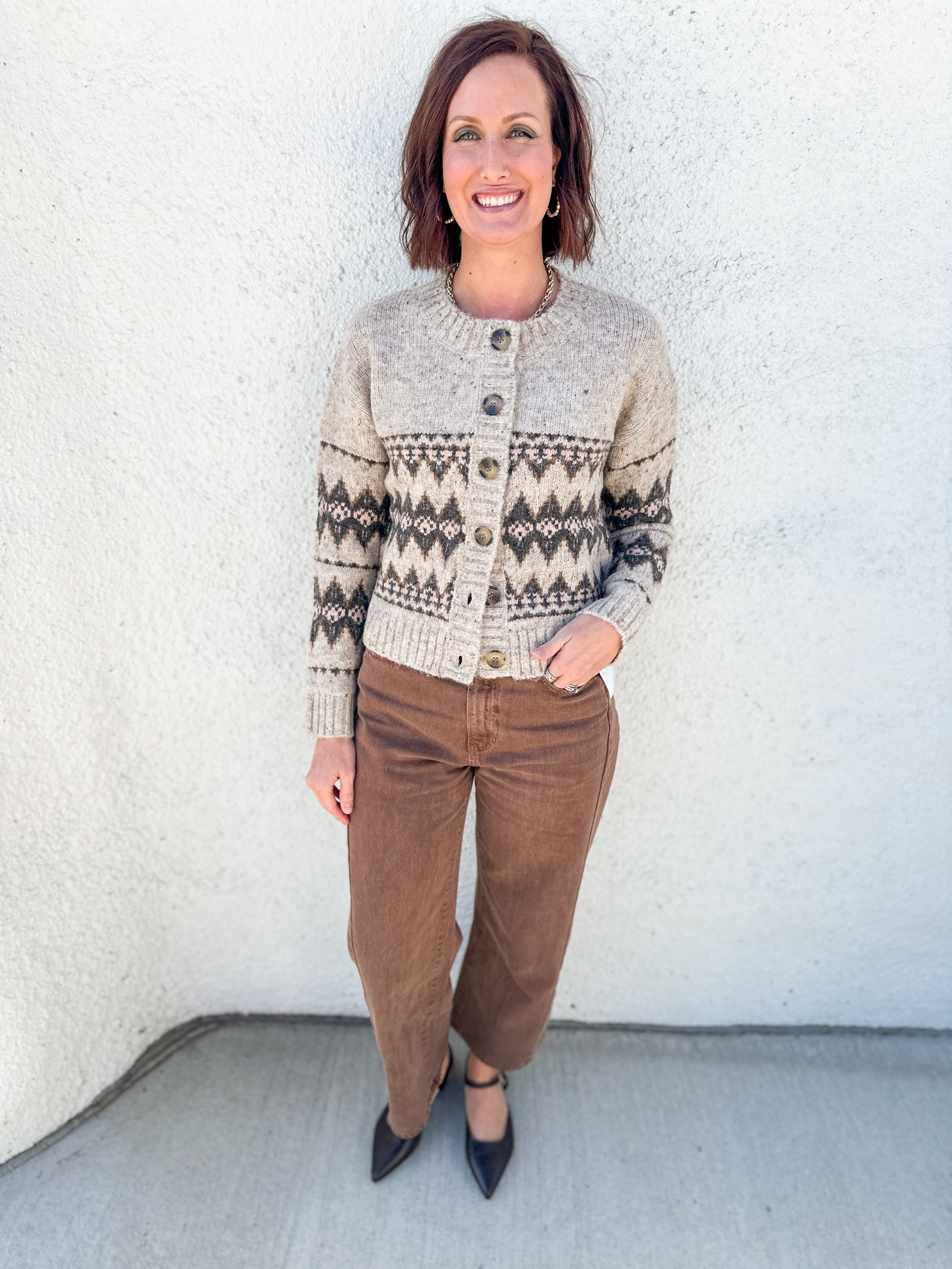 A woman stands against a white stucco wall wearing a cream and brown Fair Isle knit cardigan by Heartloom with brown barrel leg pants and mahogany brown pointed-toe flats.