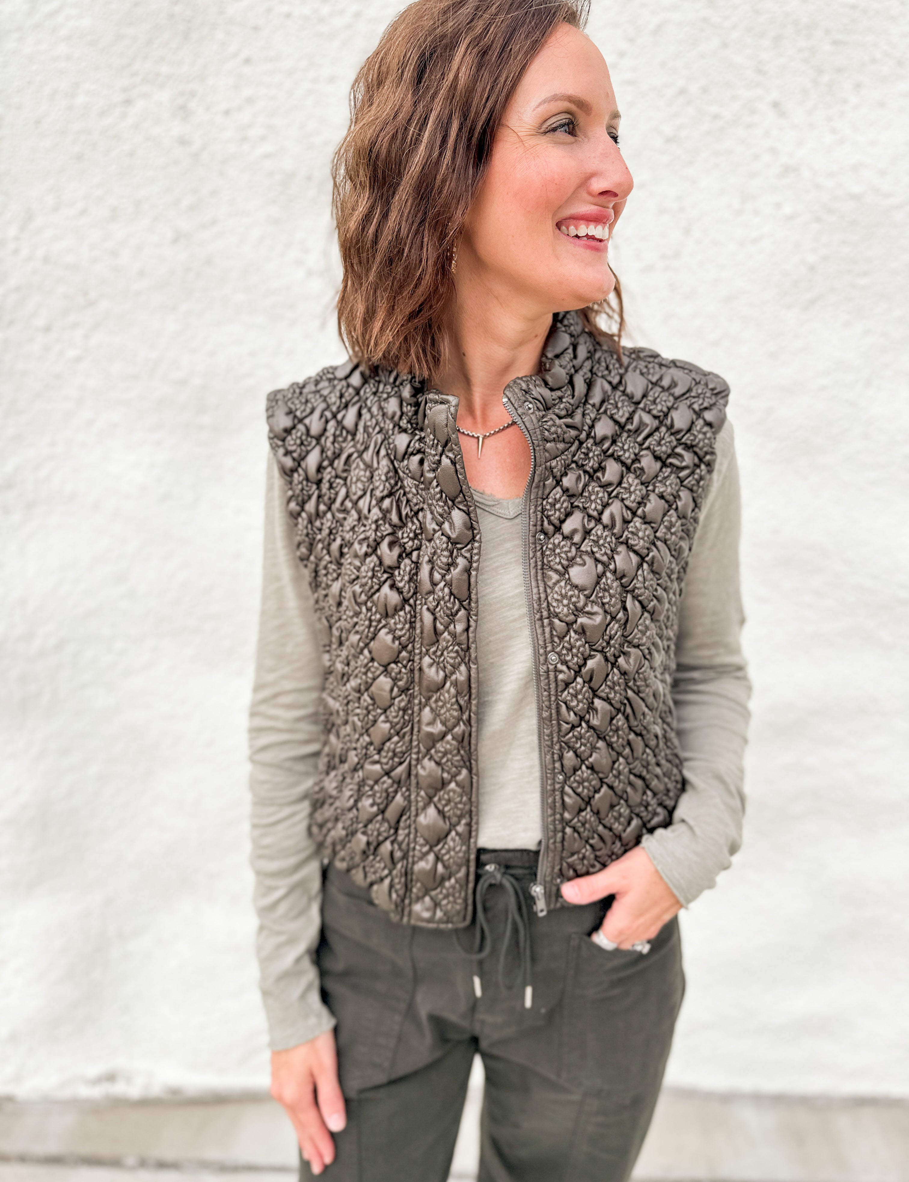 Woman wearing a quilted green vest with a faux leather look and zipper front layered over a distressed long sleeve v neck tee and green tie waist barrel jeans from Daze