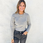 A woman wearing a light gray ribbed knit sweater and dark gray jeans, standing against a white textured wall.
