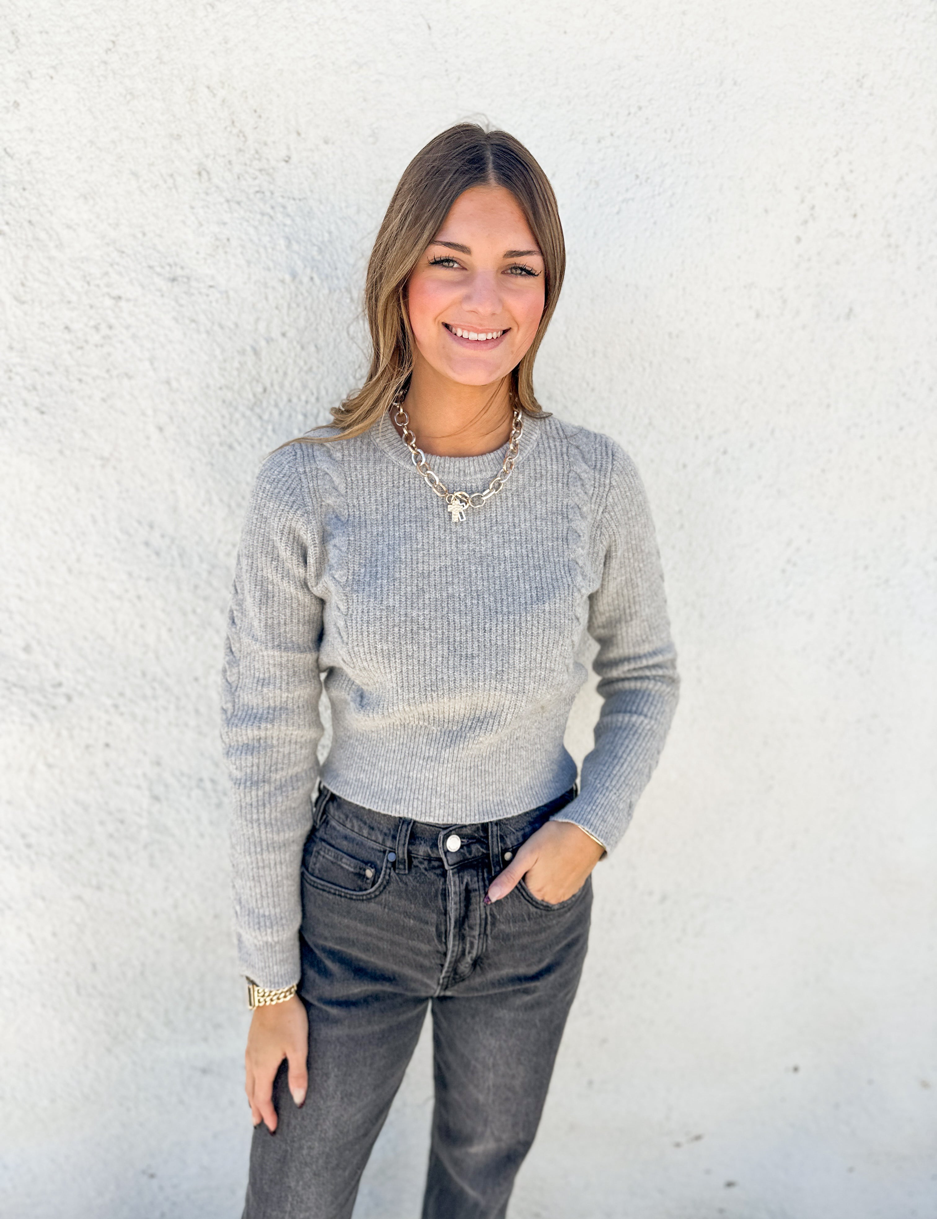 A woman wearing a light gray ribbed knit sweater and dark gray jeans, standing against a white textured wall.
