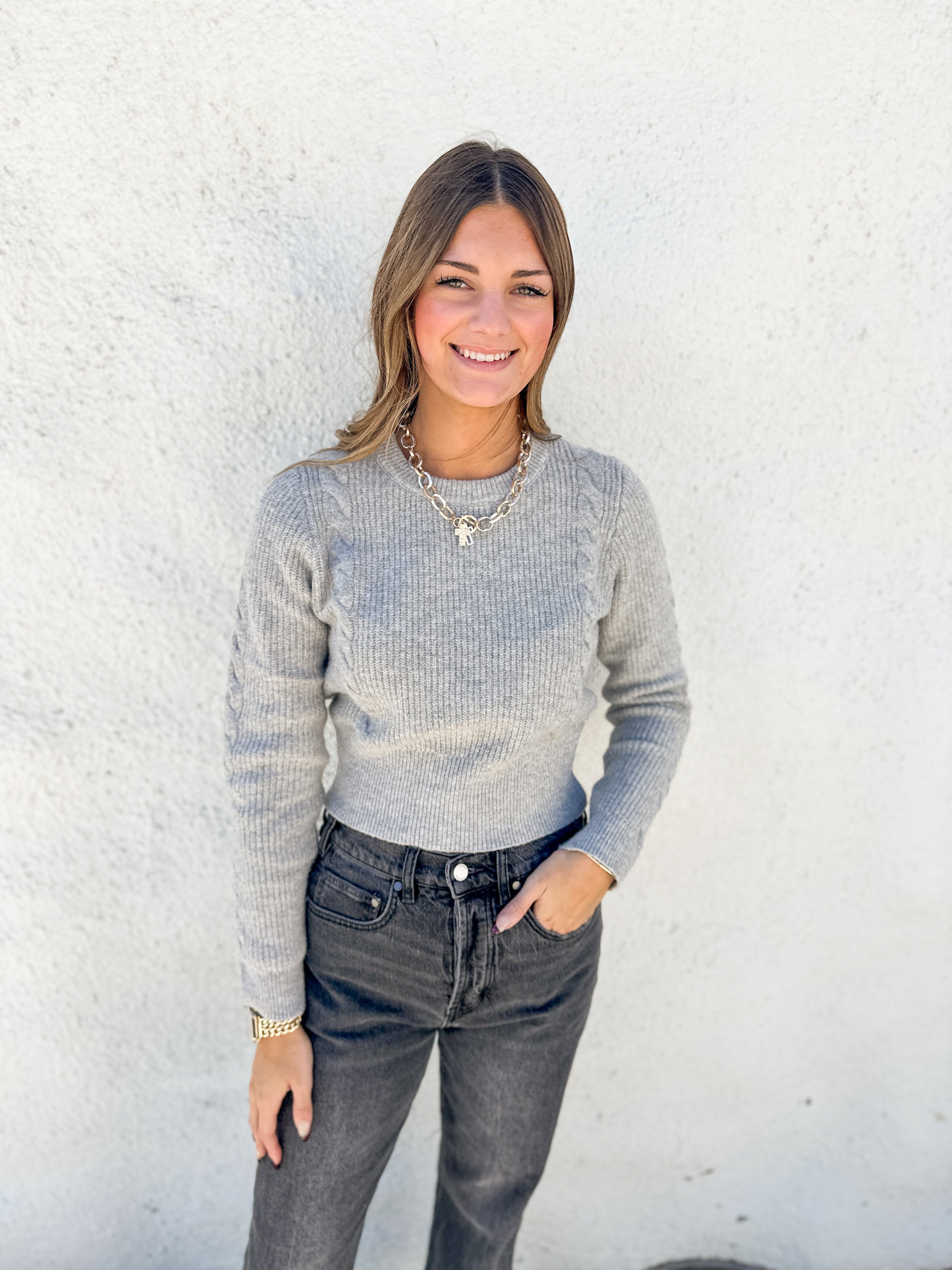 A woman wearing a light gray ribbed knit sweater and dark gray jeans, standing against a white textured wall.