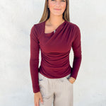 A woman modeling a long-sleeve ruched top in burgundy with a diagonal neckline, paired with light beige pants, standing against a white textured wall.