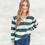 A woman wearing a soft green and cream striped long-sleeve polo shirt from Thread & Supply with blue jeans, standing against a textured white wall.