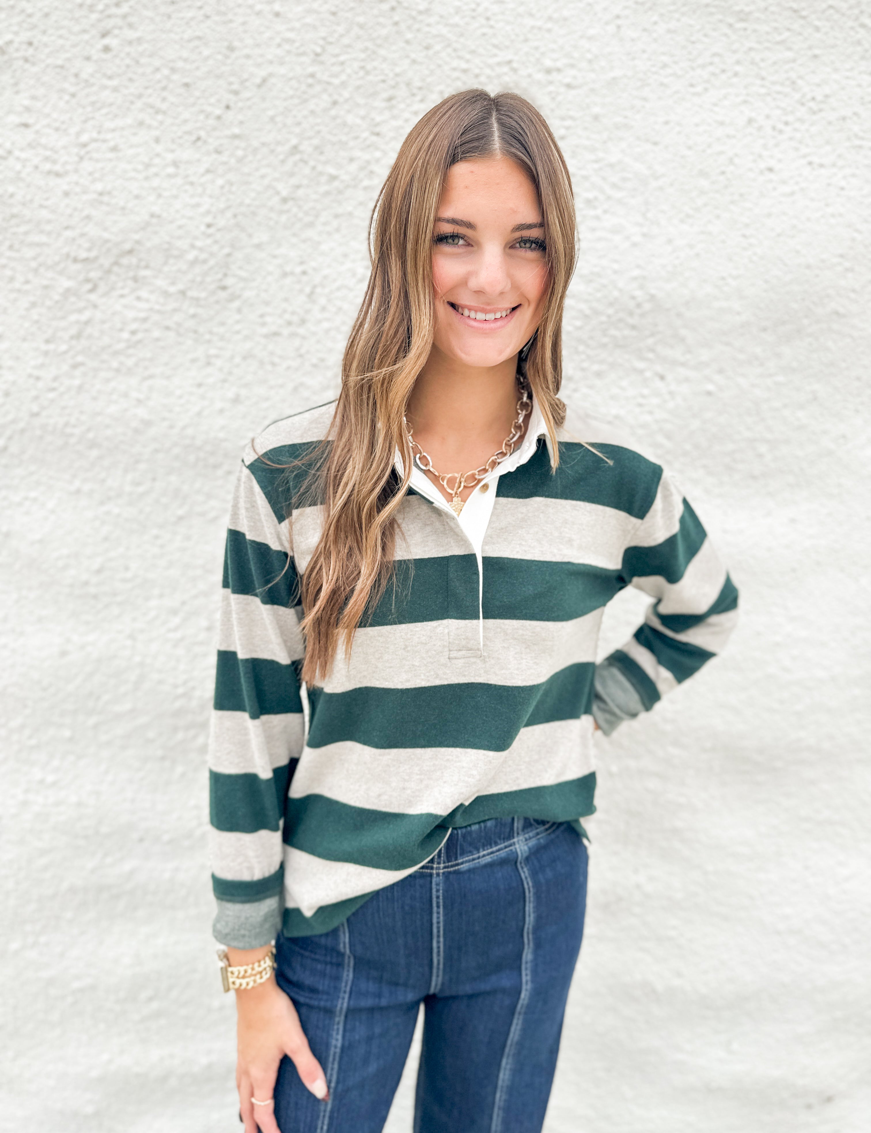 A woman wearing a soft green and cream striped long-sleeve polo shirt from Thread & Supply with blue jeans, standing against a textured white wall.