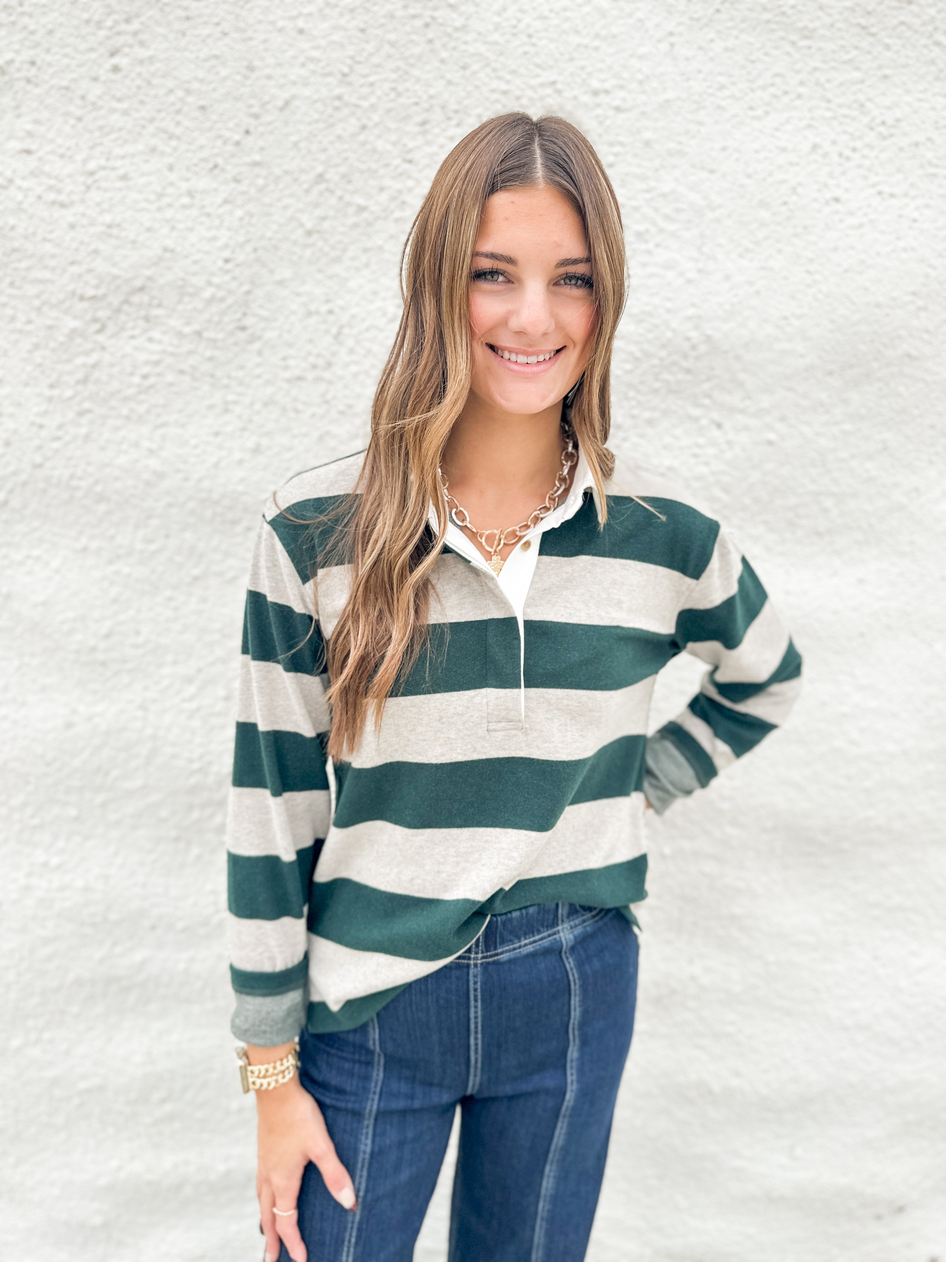 A woman wearing a soft green and cream striped long-sleeve polo shirt from Thread & Supply with blue jeans, standing against a textured white wall.