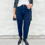 A person wearing navy blue straight-leg or relaxed-fit pants, styled with a white top, a gray plaid cardigan, and black pointed-toe ankle boots, standing against a white stucco wall.