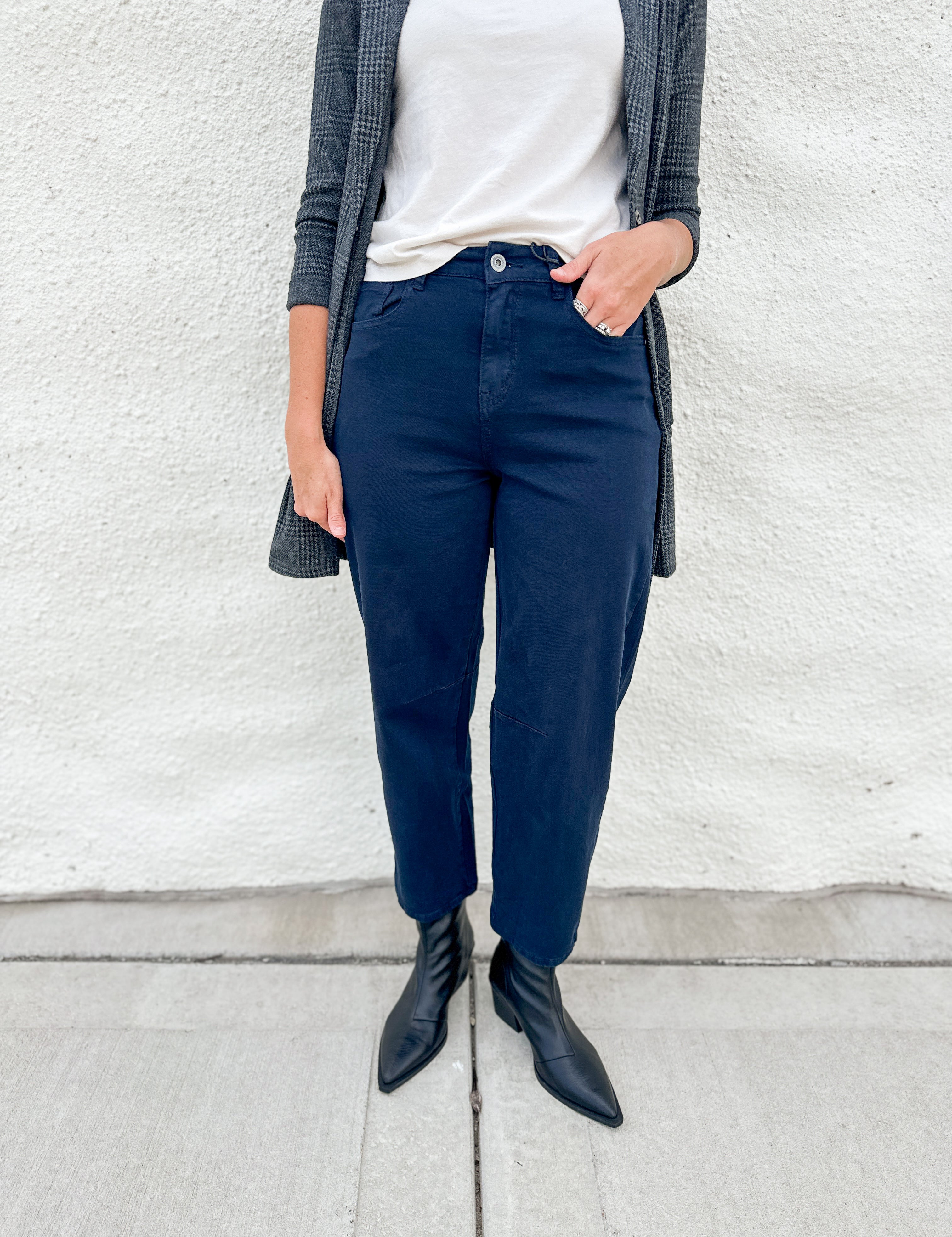 A person wearing navy blue straight-leg or relaxed-fit pants, styled with a white top, a gray plaid cardigan, and black pointed-toe ankle boots, standing against a white stucco wall.
