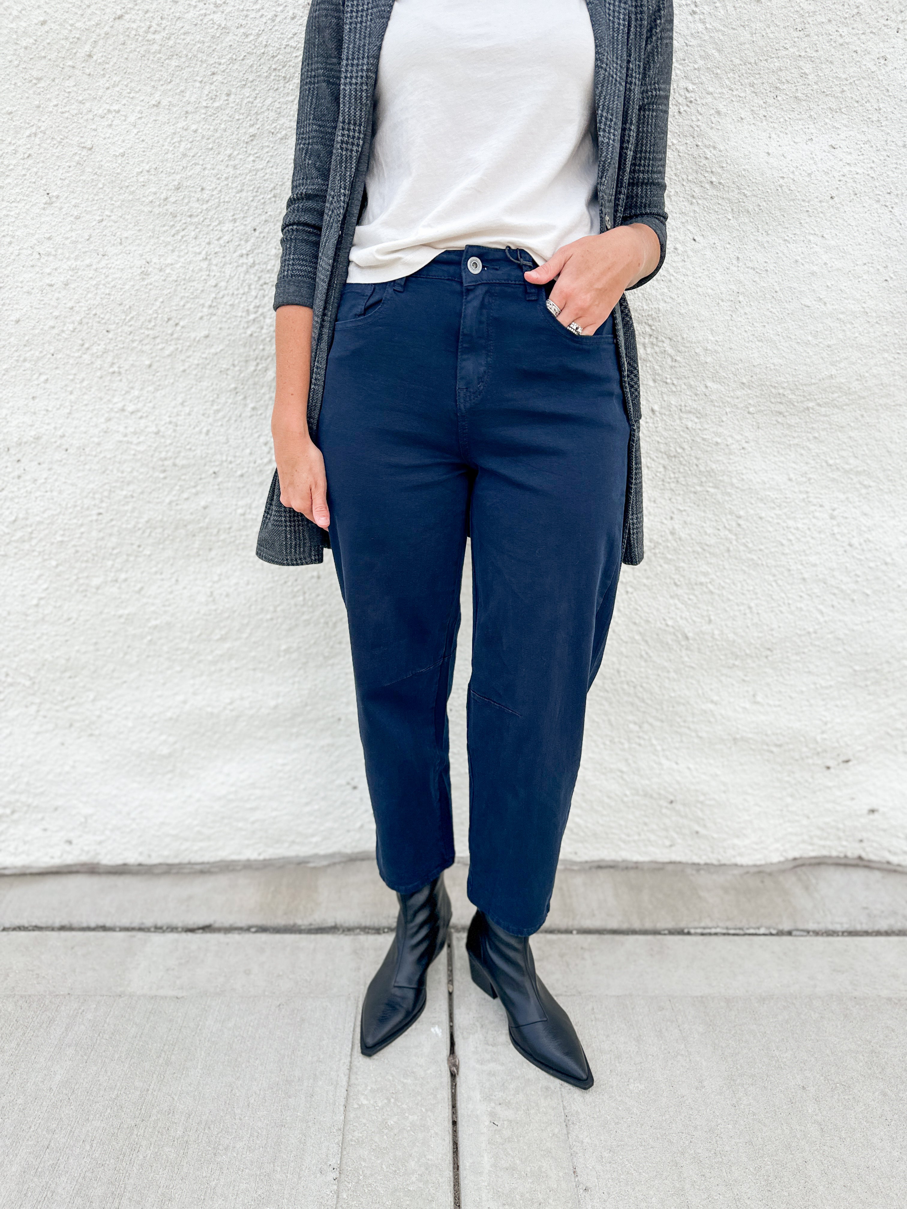 A person wearing navy blue straight-leg or relaxed-fit pants, styled with a white top, a gray plaid cardigan, and black pointed-toe ankle boots, standing against a white stucco wall.