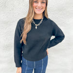 A woman with long light brown hair smiling, wearing a black wool knit crewneck sweater and blue jeans, standing against a white textured wall.