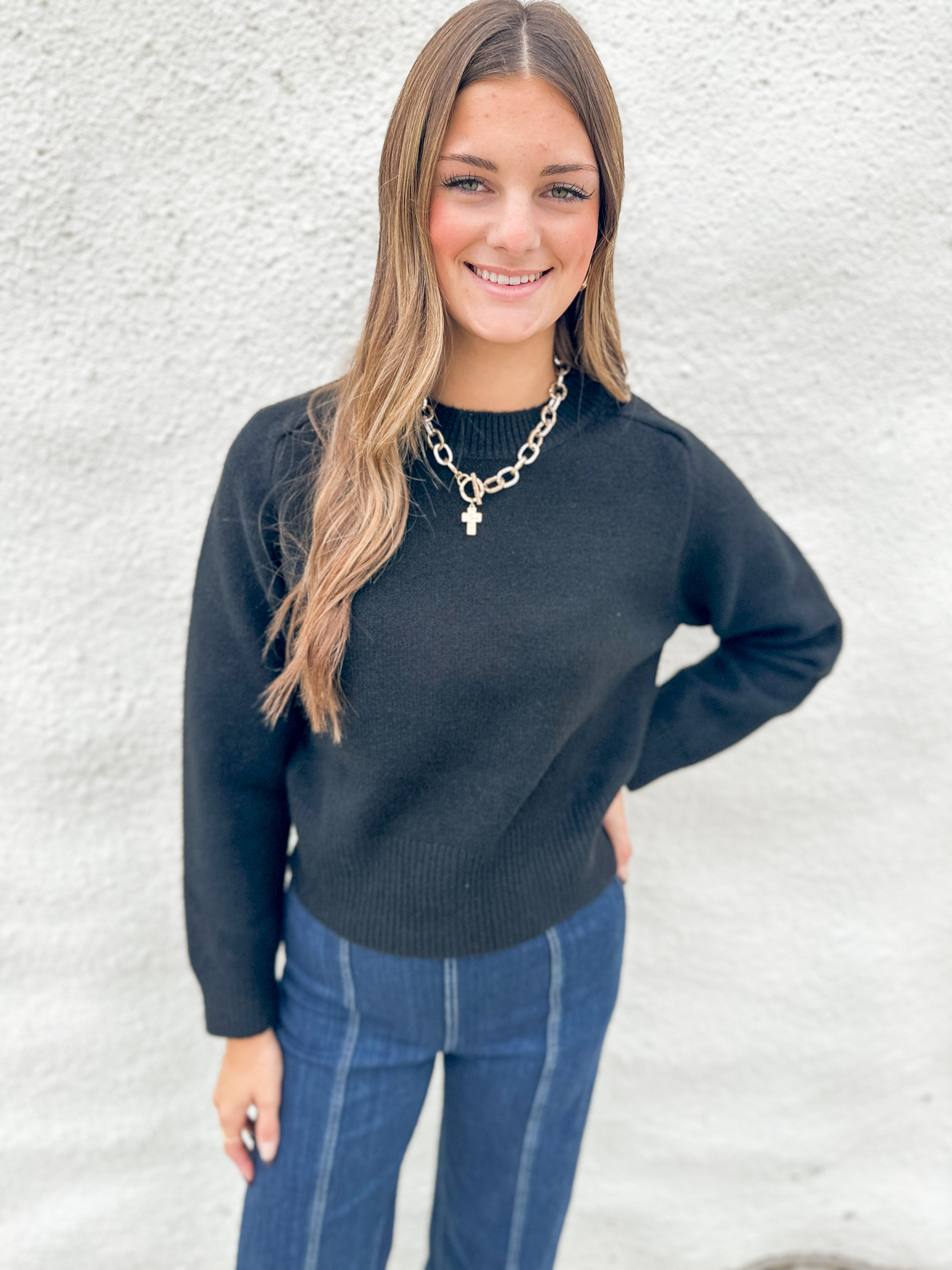 A woman with long light brown hair smiling, wearing a black wool knit crewneck sweater and blue jeans, standing against a white textured wall.