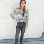 A woman stands outdoors against a white stucco wall wearing a fuzzy long-sleeve knit sweater with thin gray and white horizontal stripes, dark gray jeans, and black loafers.