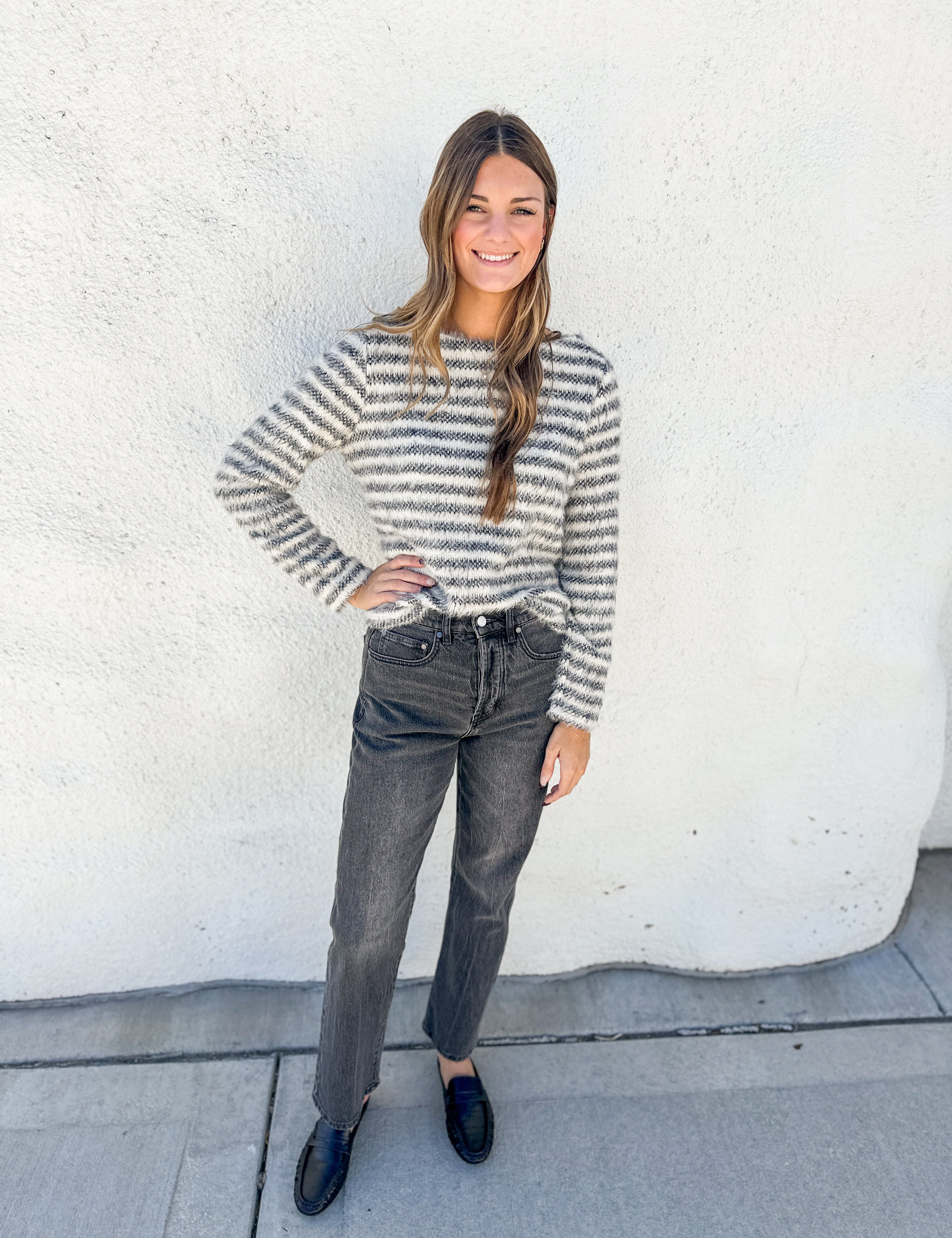 A woman stands outdoors against a white stucco wall wearing a fuzzy long-sleeve knit sweater with thin gray and white horizontal stripes, dark gray jeans, and black loafers.