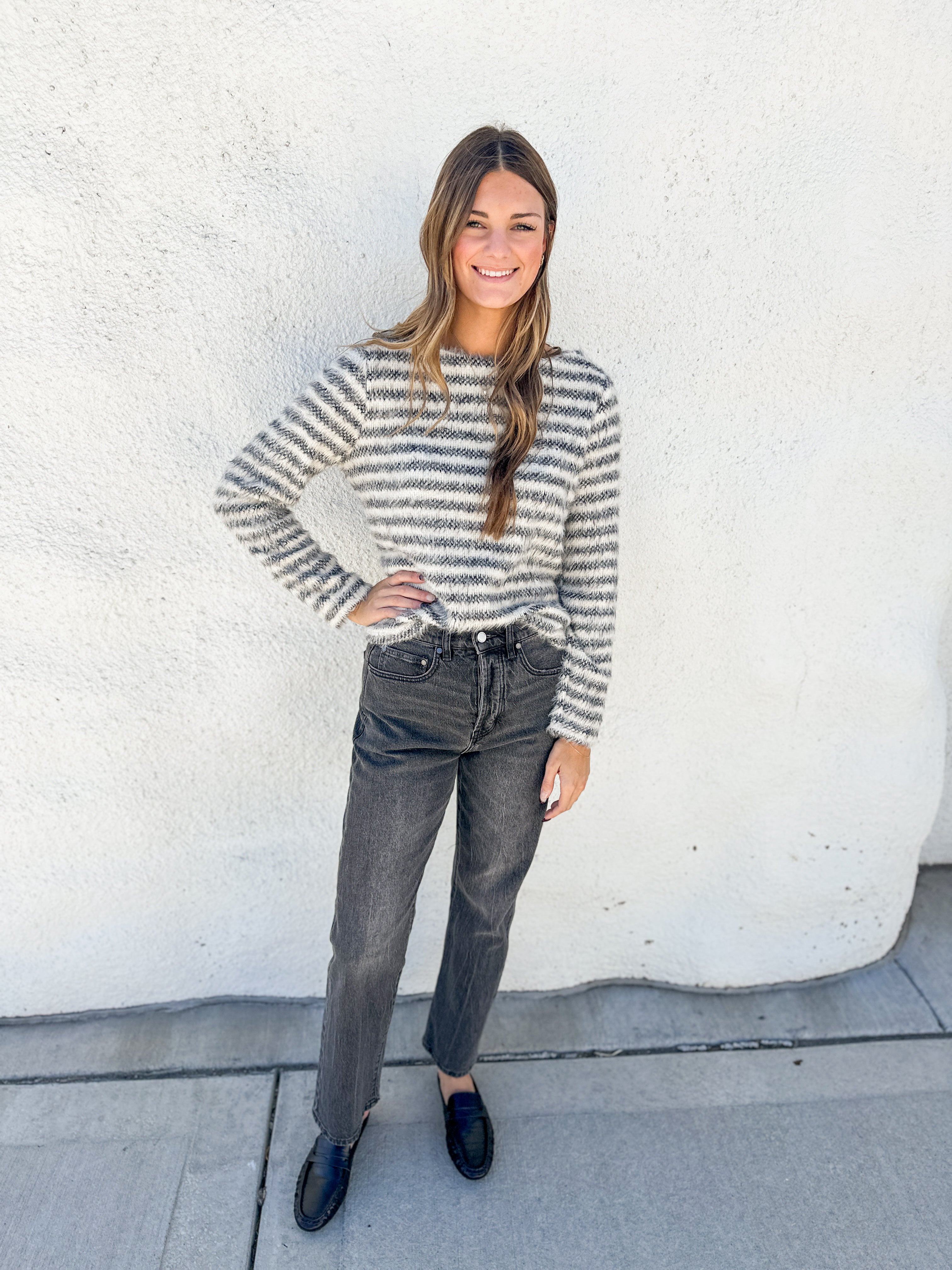 A woman stands outdoors against a white stucco wall wearing a fuzzy long-sleeve knit sweater with thin gray and white horizontal stripes, dark gray jeans, and black loafers.