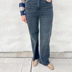 A person wearing dark blue/black wide-leg jeans with raw hem, paired with a blue and beige striped long-sleeve top and beige driving moccasins with gold stud detailing, standing against a white textured wall.