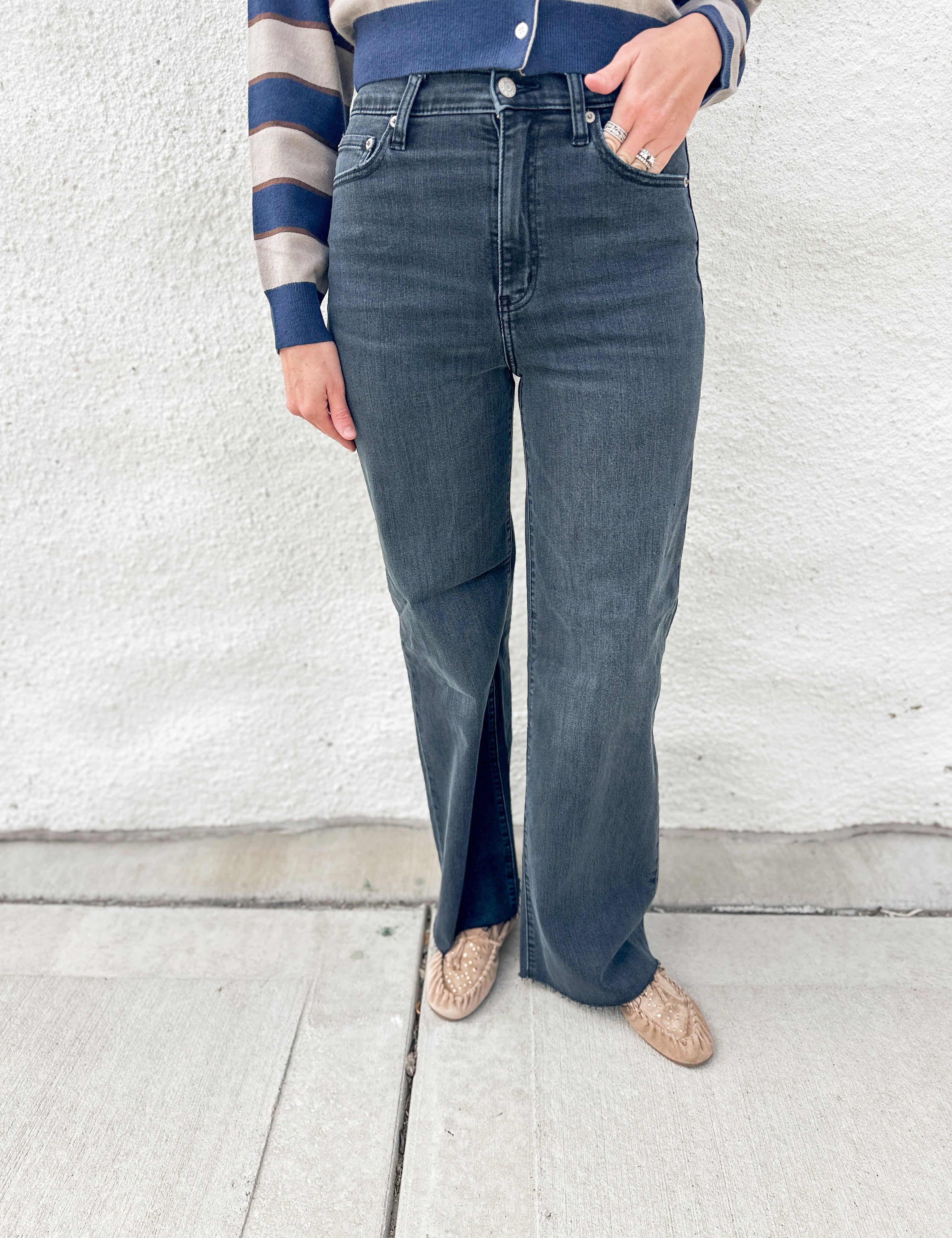 A person wearing dark blue/black wide-leg jeans with raw hem, paired with a blue and beige striped long-sleeve top and beige driving moccasins with gold stud detailing, standing against a white textured wall.
