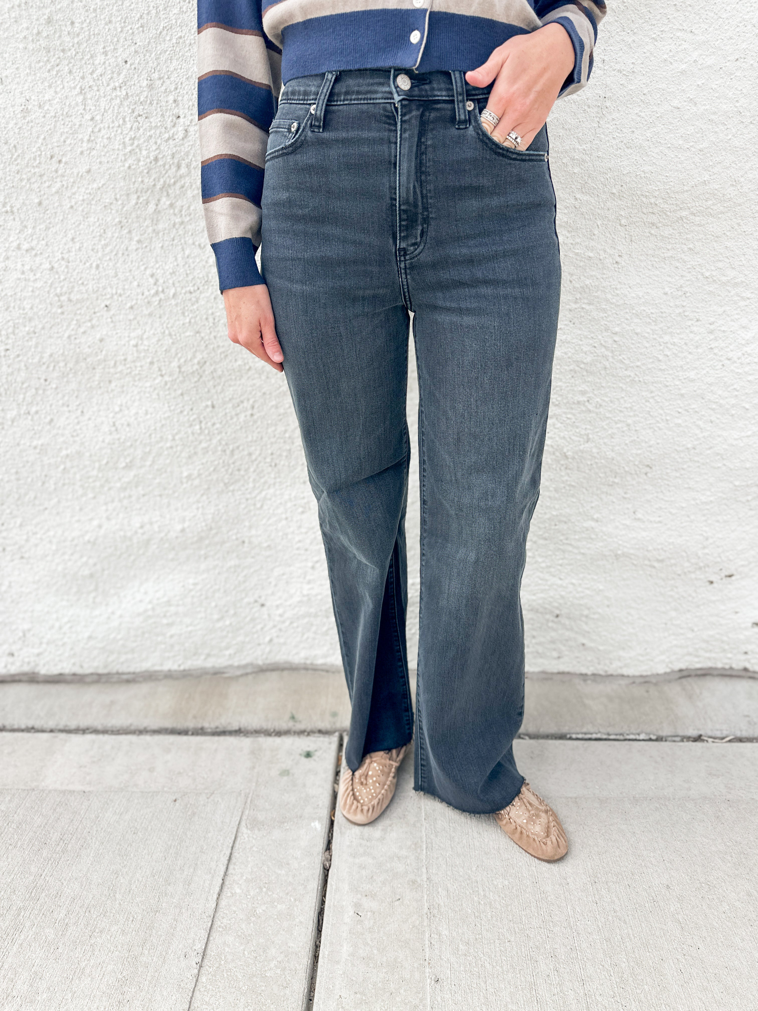 A person wearing dark blue/black wide-leg jeans with raw hem, paired with a blue and beige striped long-sleeve top and beige driving moccasins with gold stud detailing, standing against a white textured wall.