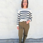 A woman stands against a white stucco wall wearing a cream knit sweater with black horizontal stripes, olive green pants, and black shoes.