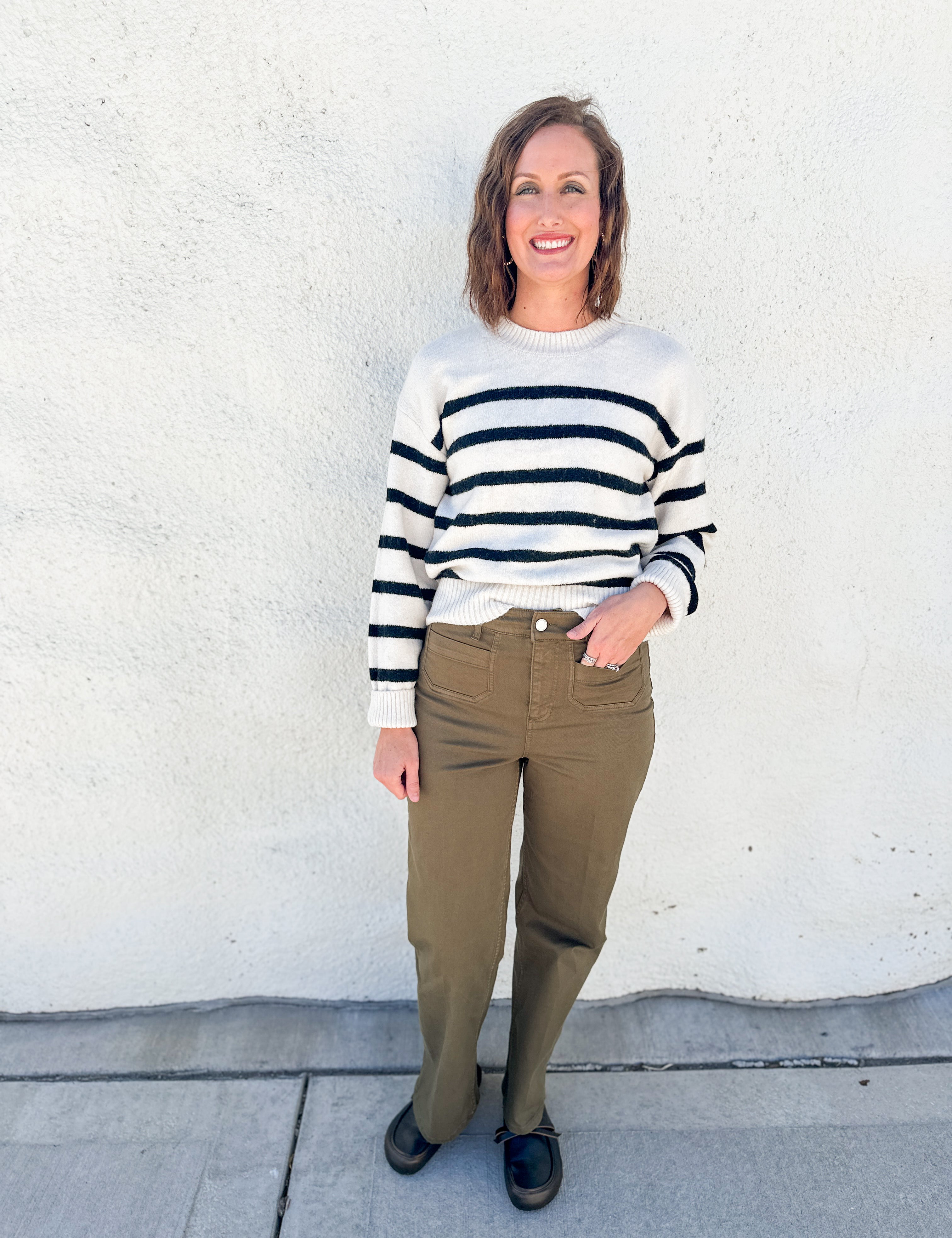 A woman stands against a white stucco wall wearing a cream knit sweater with black horizontal stripes, olive green pants, and black shoes.