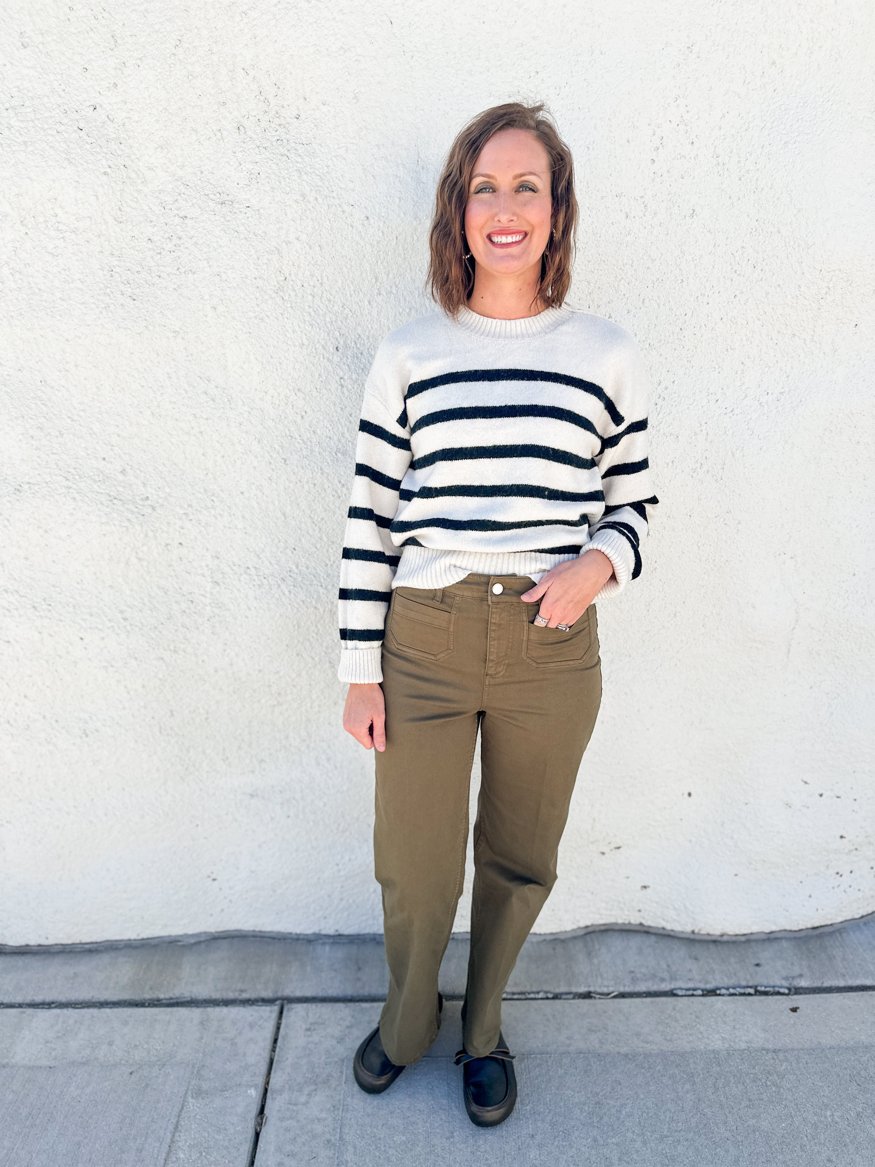 A woman stands against a white stucco wall wearing a cream knit sweater with black horizontal stripes, olive green pants, and black shoes.