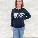 A woman wearing a black crewneck sweater with 'HOCKEY' printed in bold white letters across the chest, paired with blue jeans, standing against a white textured wall.