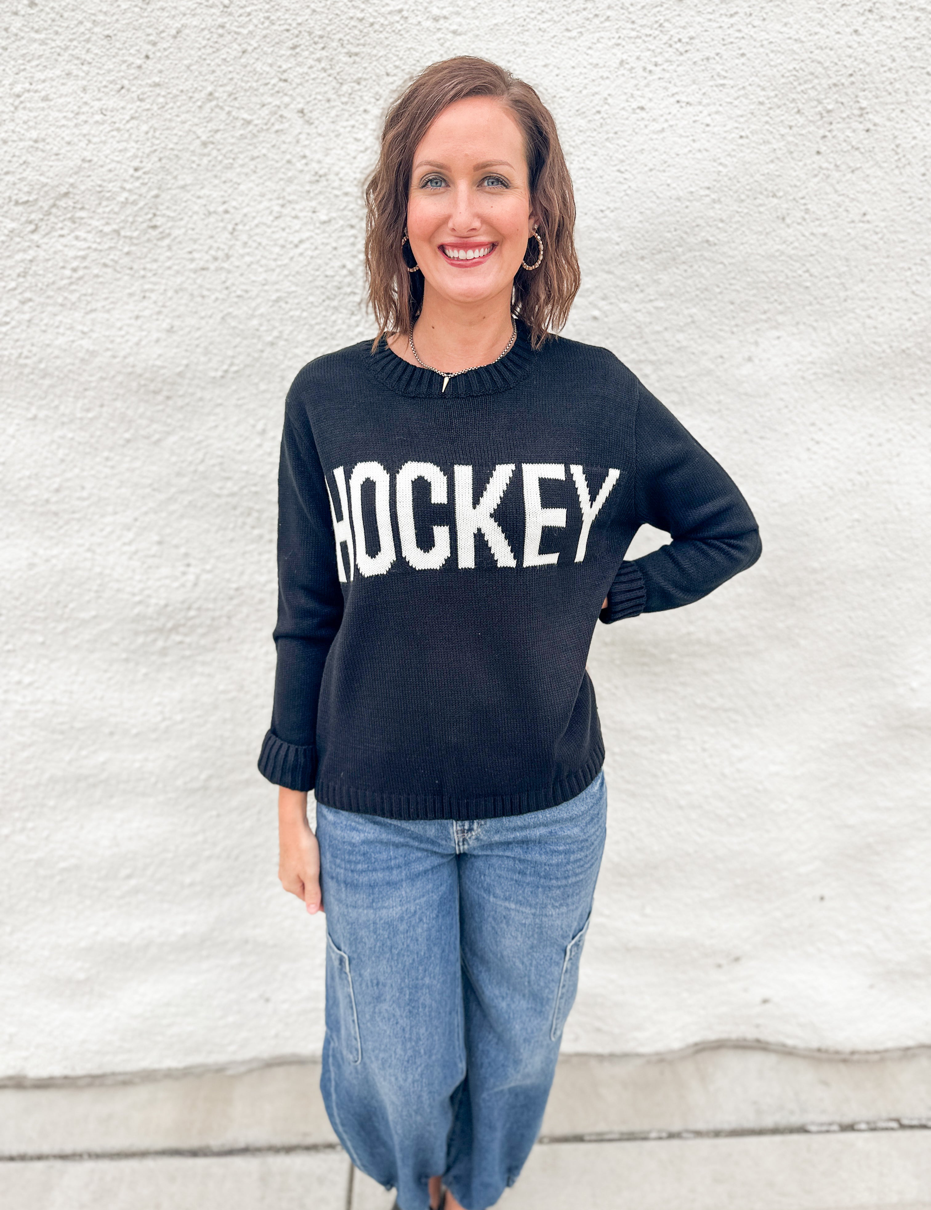 A woman wearing a black crewneck sweater with 'HOCKEY' printed in bold white letters across the chest, paired with blue jeans, standing against a white textured wall.