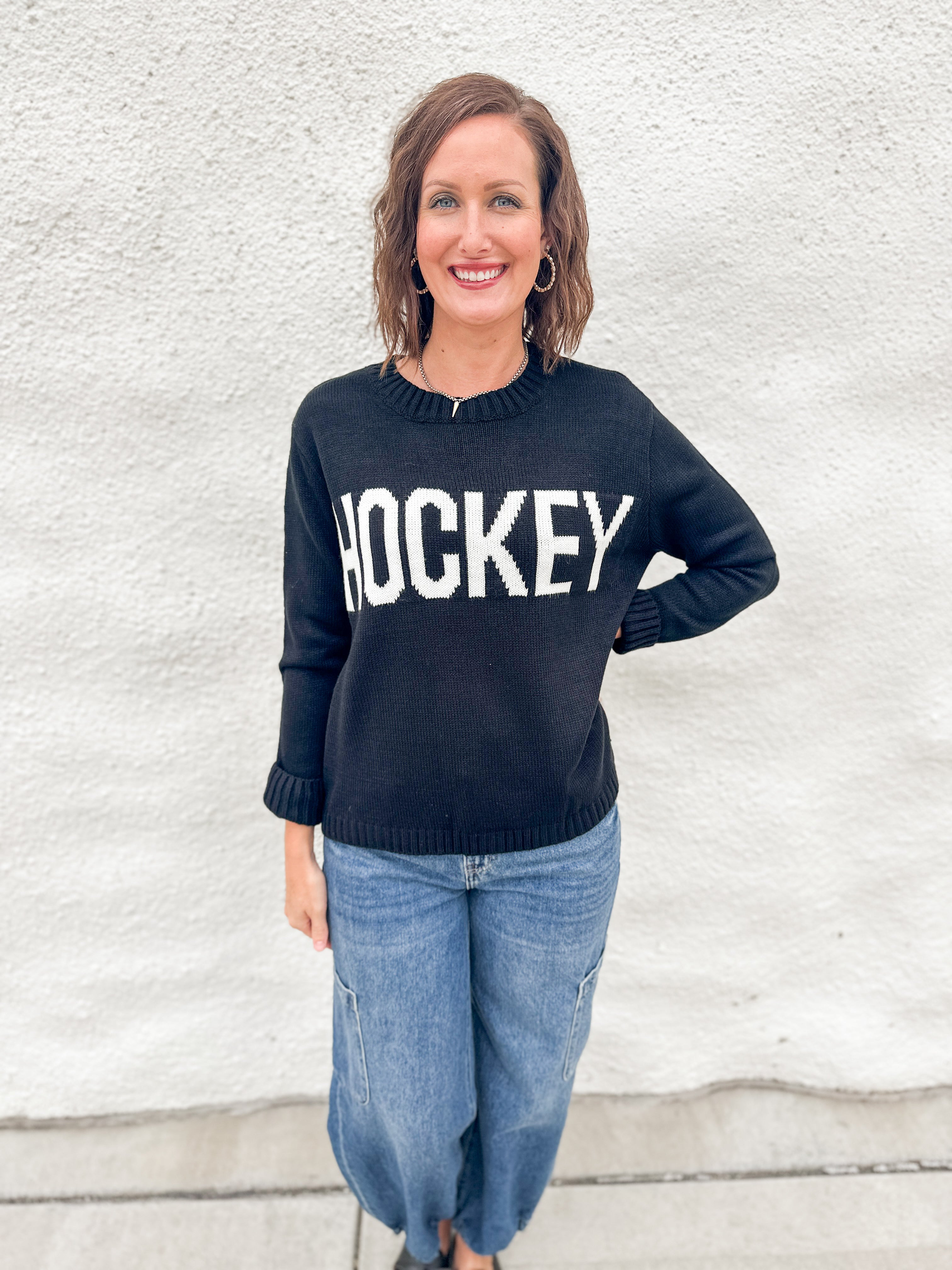 A woman wearing a black crewneck sweater with 'HOCKEY' printed in bold white letters across the chest, paired with blue jeans, standing against a white textured wall.
