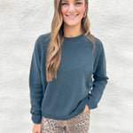 A woman wearing a washed black crewneck fleece lined sweatshirt from Daydreamer and leopard print pants, standing against a textured white wall.