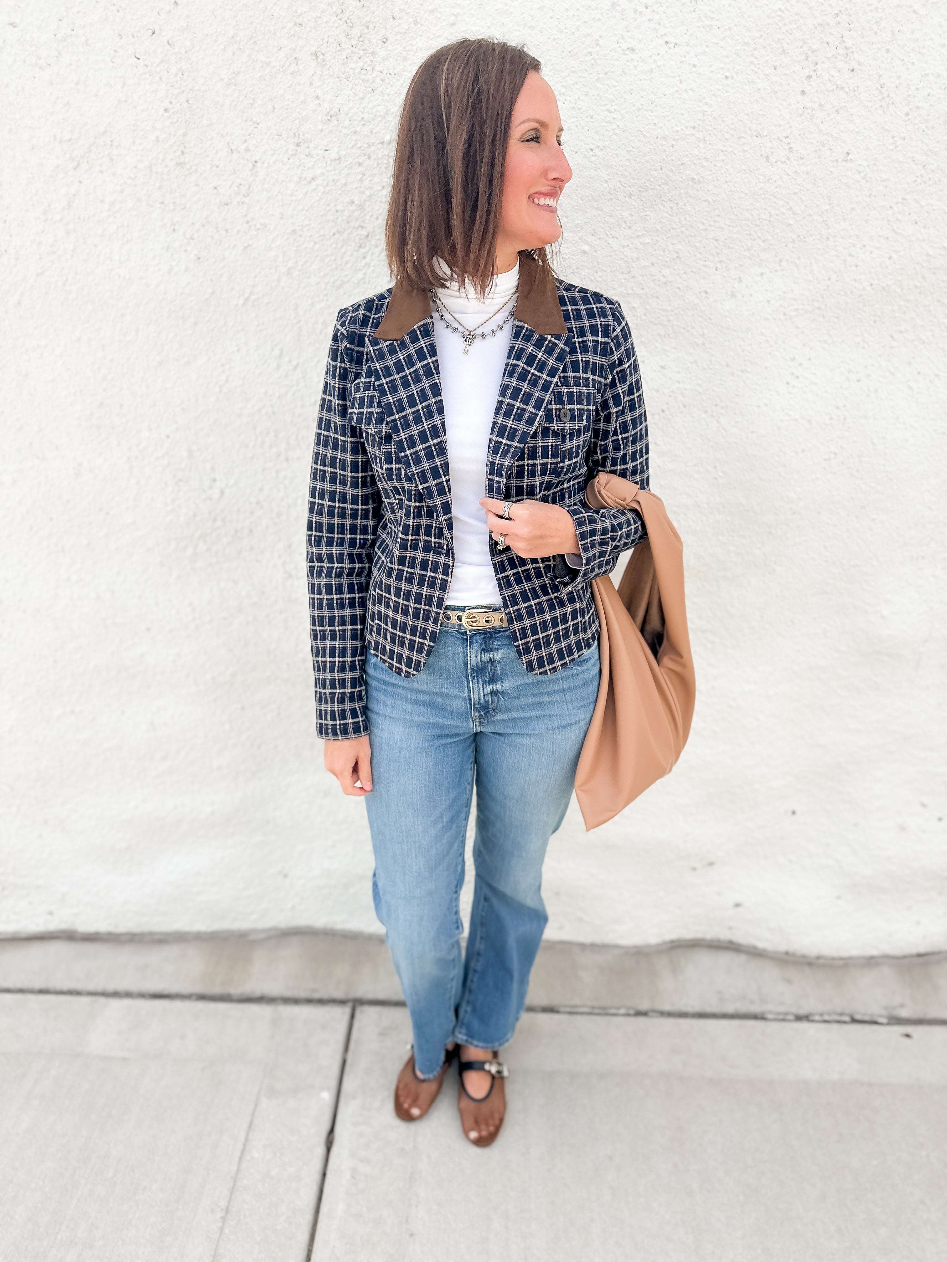 A woman stands against a white stucco wall wearing a navy and beige plaid blazer with brown suede lapels, a white turtleneck, blue jeans, and brown mesh mary jane flats, while carrying a tan shoulder bag