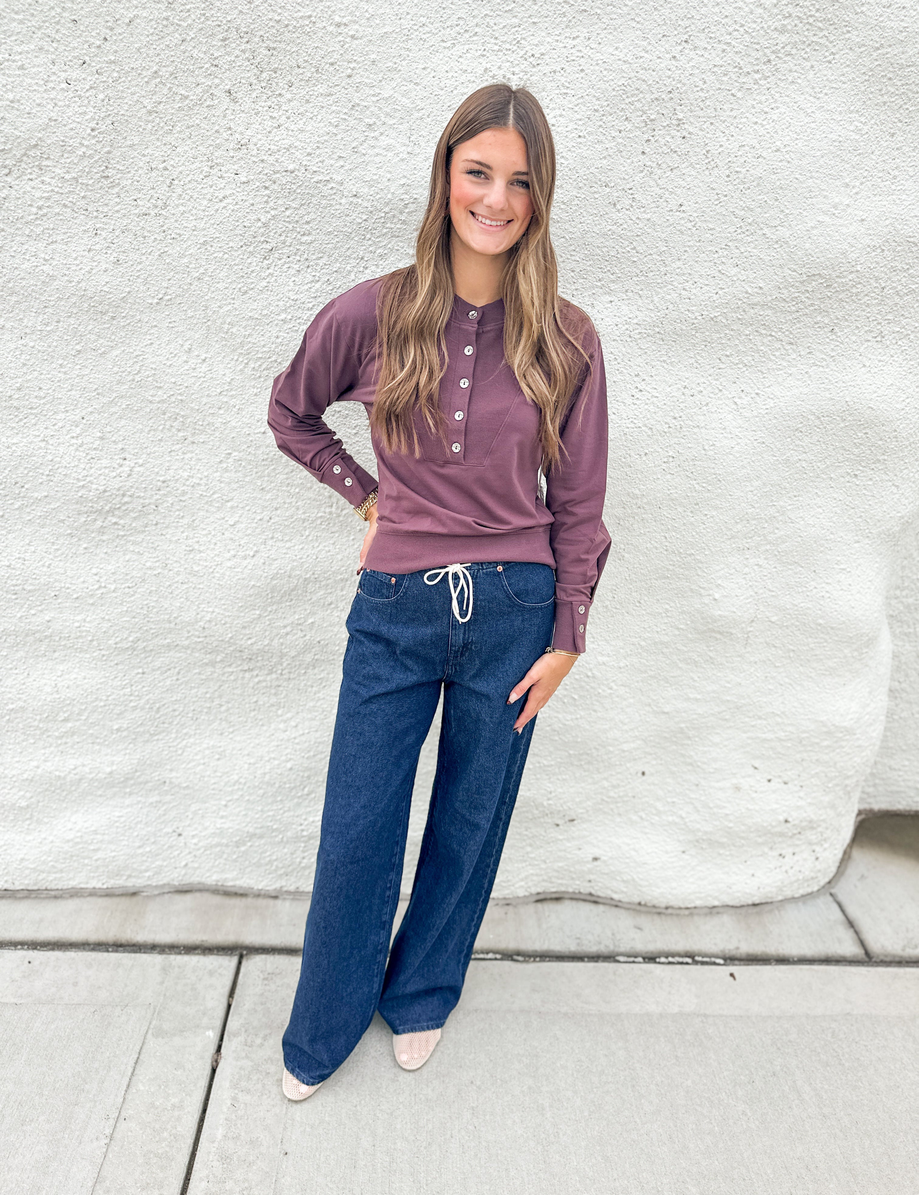 Smiling model wearing a full length wide leg denim with cream drawstring waist tie from Daze denim with a purple long sleeve top with front and sleeve button details.